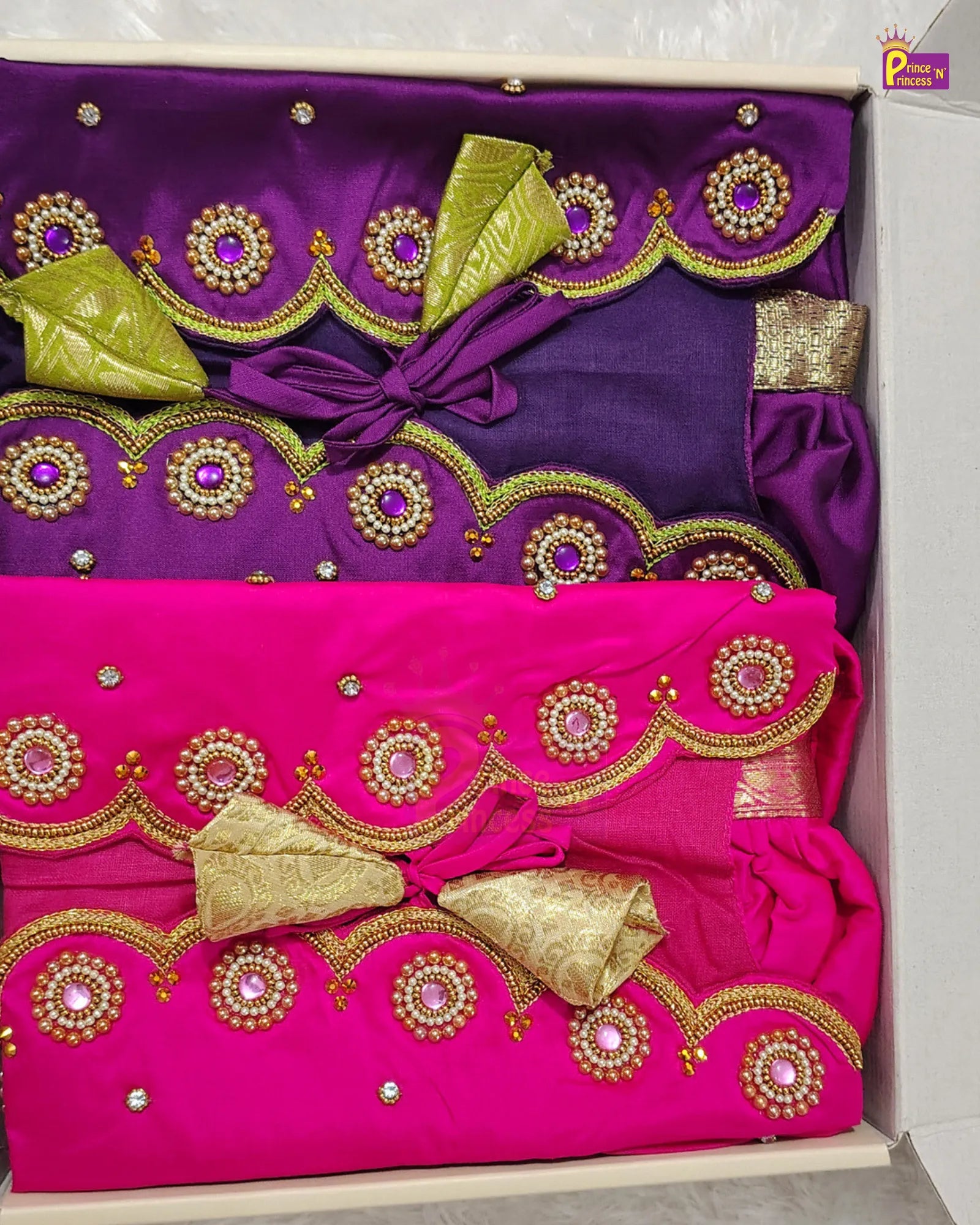 Two folded traditional Indian garments stacked inside a light-colored box. The top garment is deep purple with gold and white embroidered scalloped borders and a gold bow.