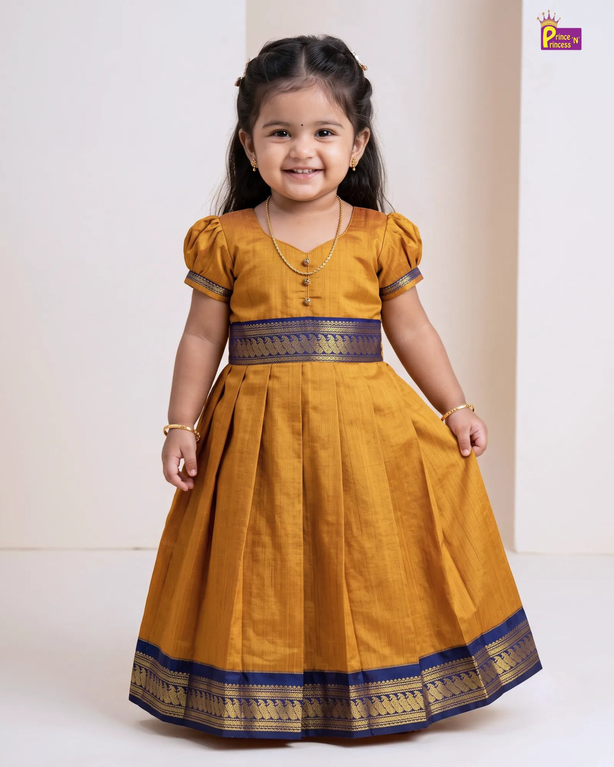 A young girl wearing a mustard yellow traditional dress with a navy blue and gold patterned waistband and hem, standing against a plain off-white background.