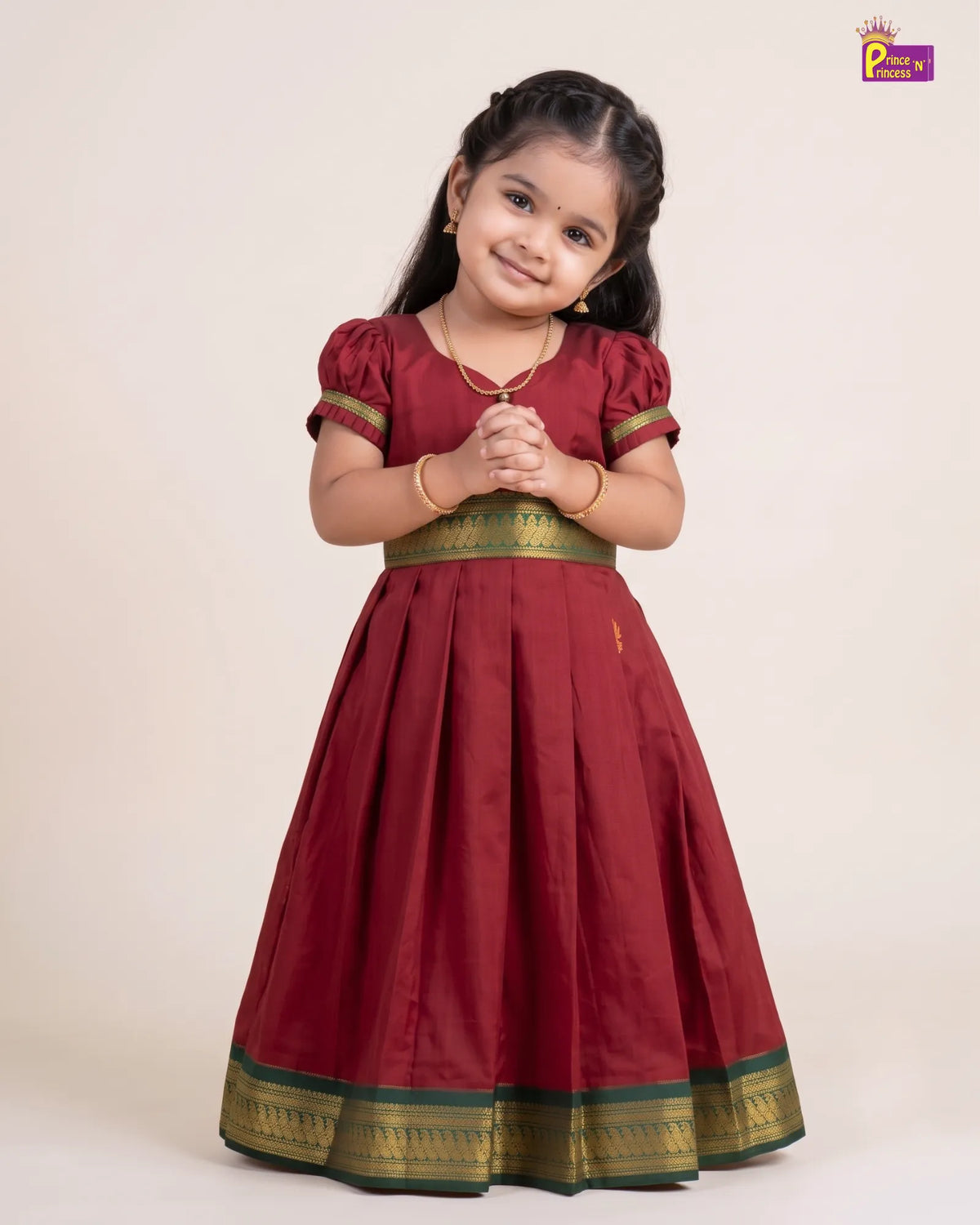 A young girl modeling a traditional Indian dress, featuring a deep maroon skirt with a gold and green border, short puff sleeves, and a matching waistband. She is standing against a plain beige background with her hands clasped together.