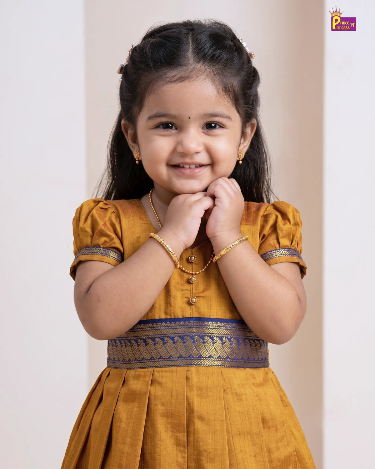 A young girl with dark hair styled in half-ponchos, wearing a mustard yellow traditional dress with gold and blue patterns, posing with her hands clasped together. She is wearing gold jewelry, including a necklace and bracelets, against a soft beige background.