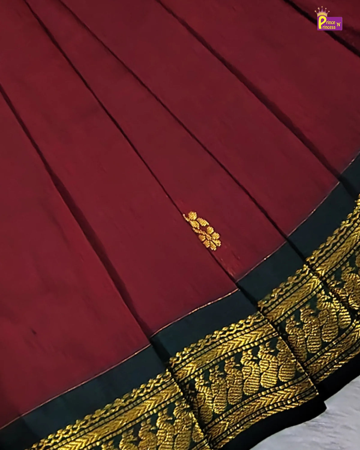 A close-up of a deep red fabric with pleated texture, featuring a small gold floral motif and a wide black and gold ornate border.