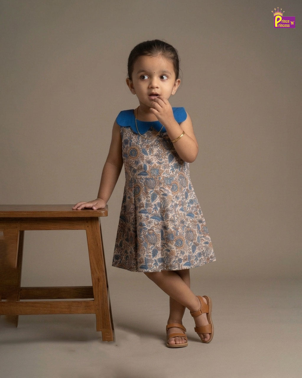 Young girl in a floral dress standing next to a wooden table against a plain background - Prince N Princess