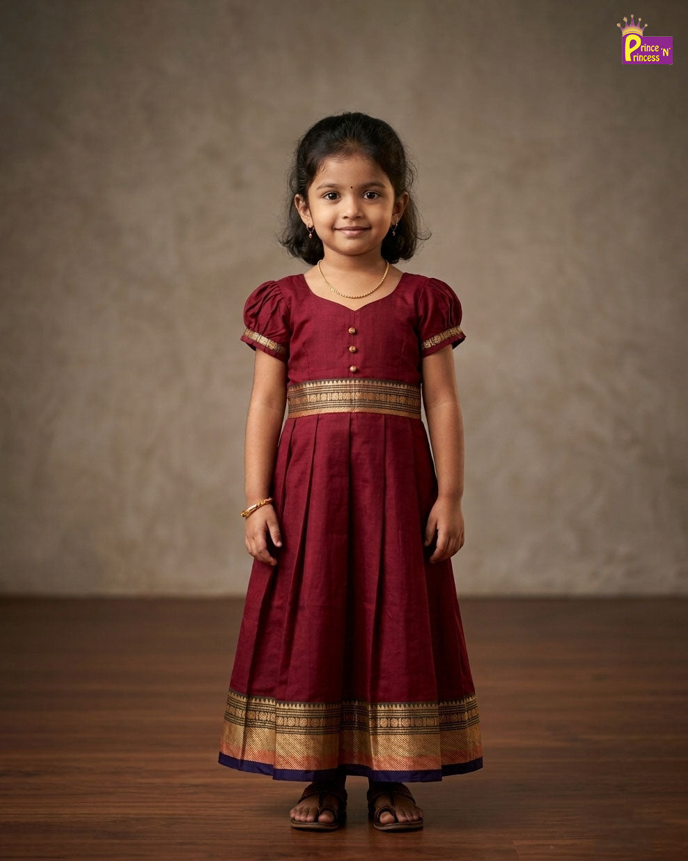 Young girl wearing a maroon traditional dress with gold accents against a plain background - Prince N Princess