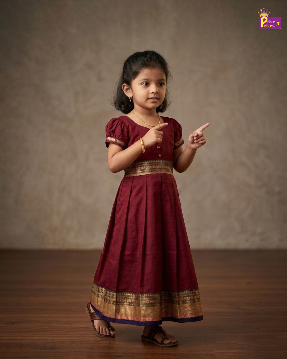 Young girl in a maroon traditional dress pointing with both hands against a plain background - Prince N Princess