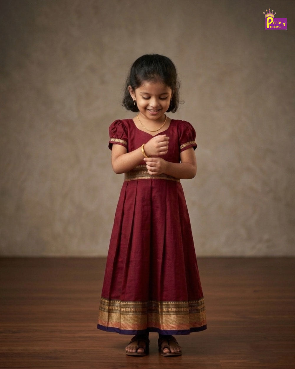 Young girl wearing a maroon traditional outfit with gold details on a wooden floor. - Prince N Princess
