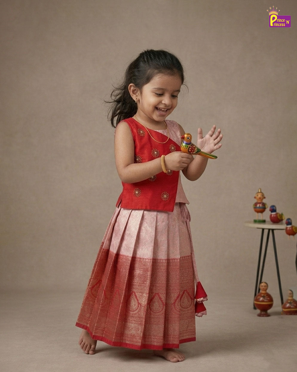Young girl in a red and pink traditional outfit holding a small toy, with a neutral background. - Prince N Princess