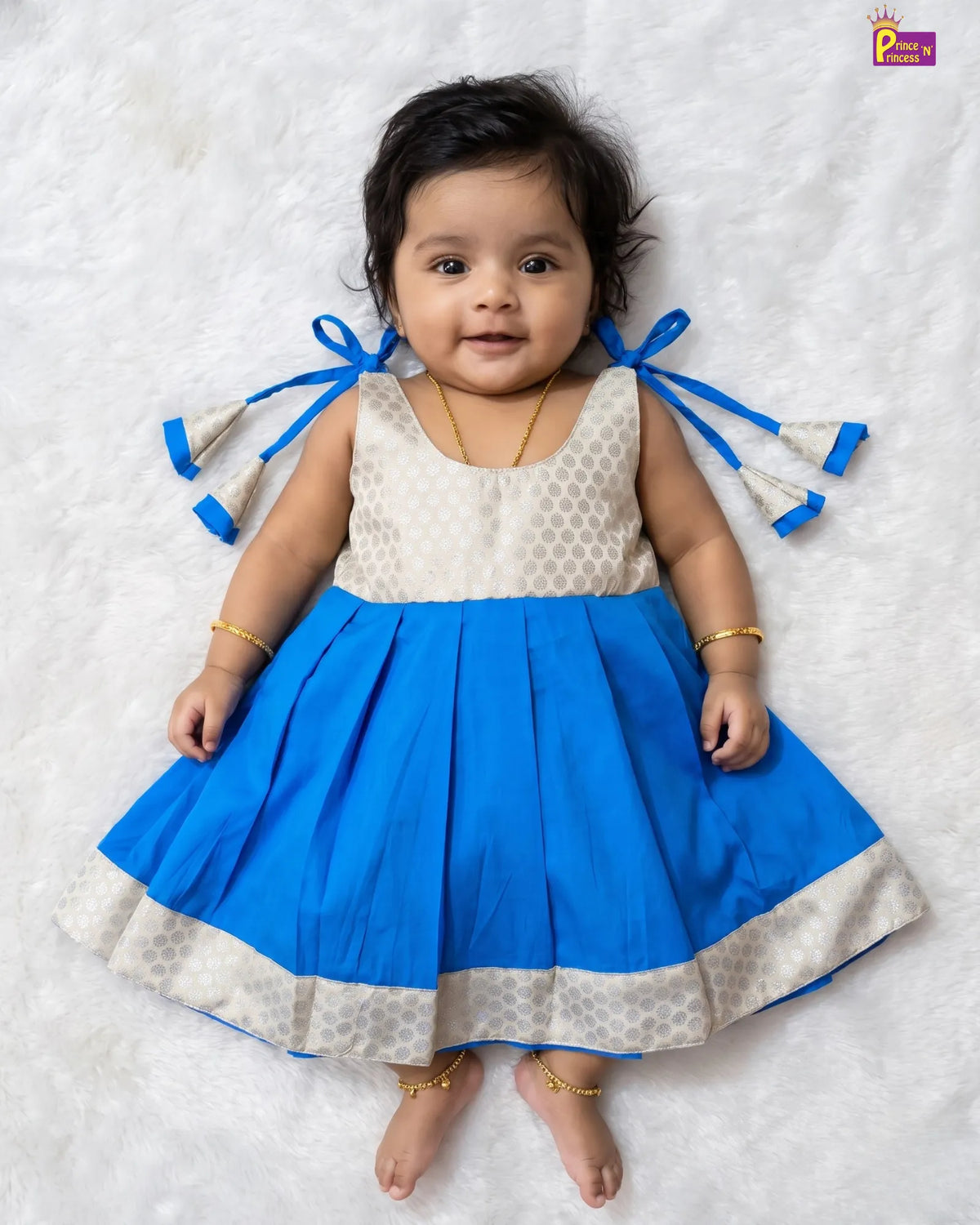A baby girl wearing a sleeveless blue and cream dress with patterned details, lying on a white textured blanket. The dress features a solid blue pleated skirt with a cream patterned hem and matching blue ribbon bows on the back.