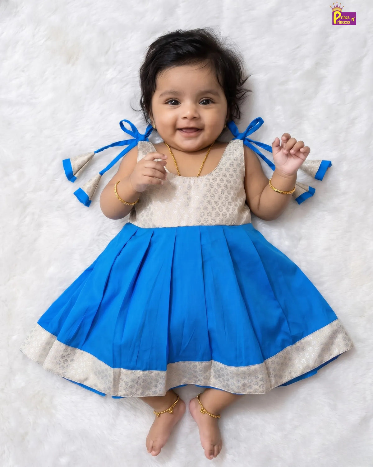 A baby girl lying on a white textured blanket, wearing a sleeveless blue and cream dress with blue ribbon bows on the sides. She is wearing gold jewelry, including a necklace, bracelets, and anklets.