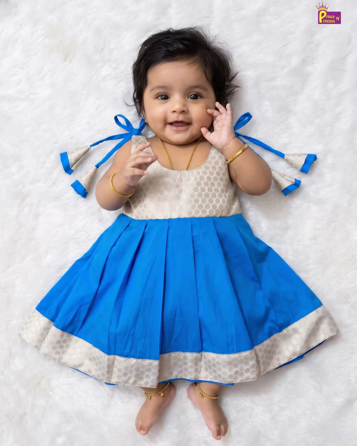 A baby girl wearing a bright blue and cream patterned dress, lying on a white textured blanket. The dress has a cream patterned bodice, a pleated blue skirt, and blue ribbon ties on the sleeves.