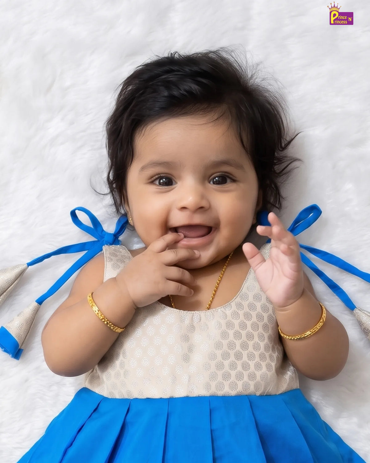 A smiling baby girl lying on a white textured blanket, wearing a cream and blue dress with gold jewelry.