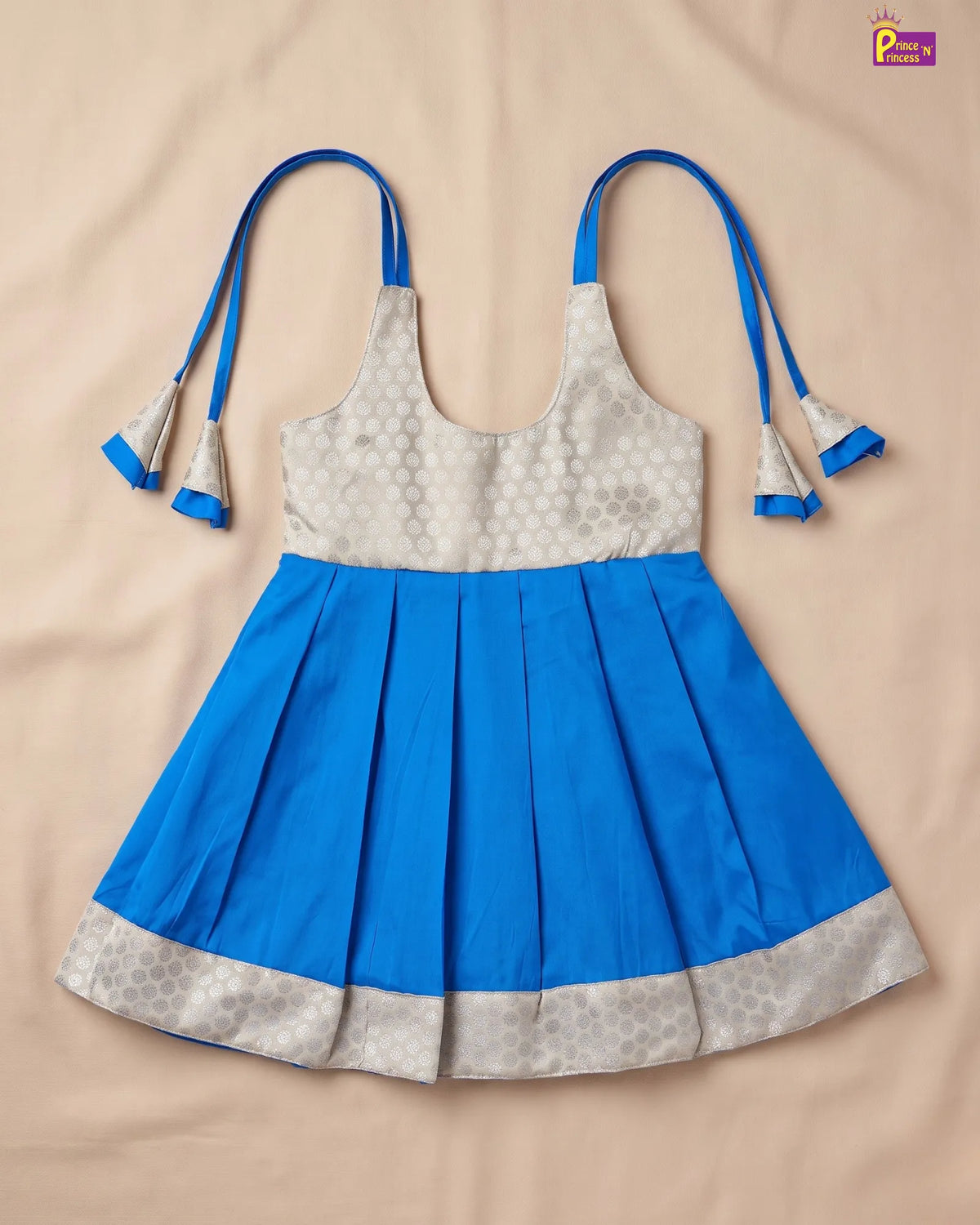A sleeveless children's dress laid flat on a beige fabric background. The dress has a bright blue pleated skirt and a light beige/cream patterned bodice with blue tie straps and tassels.