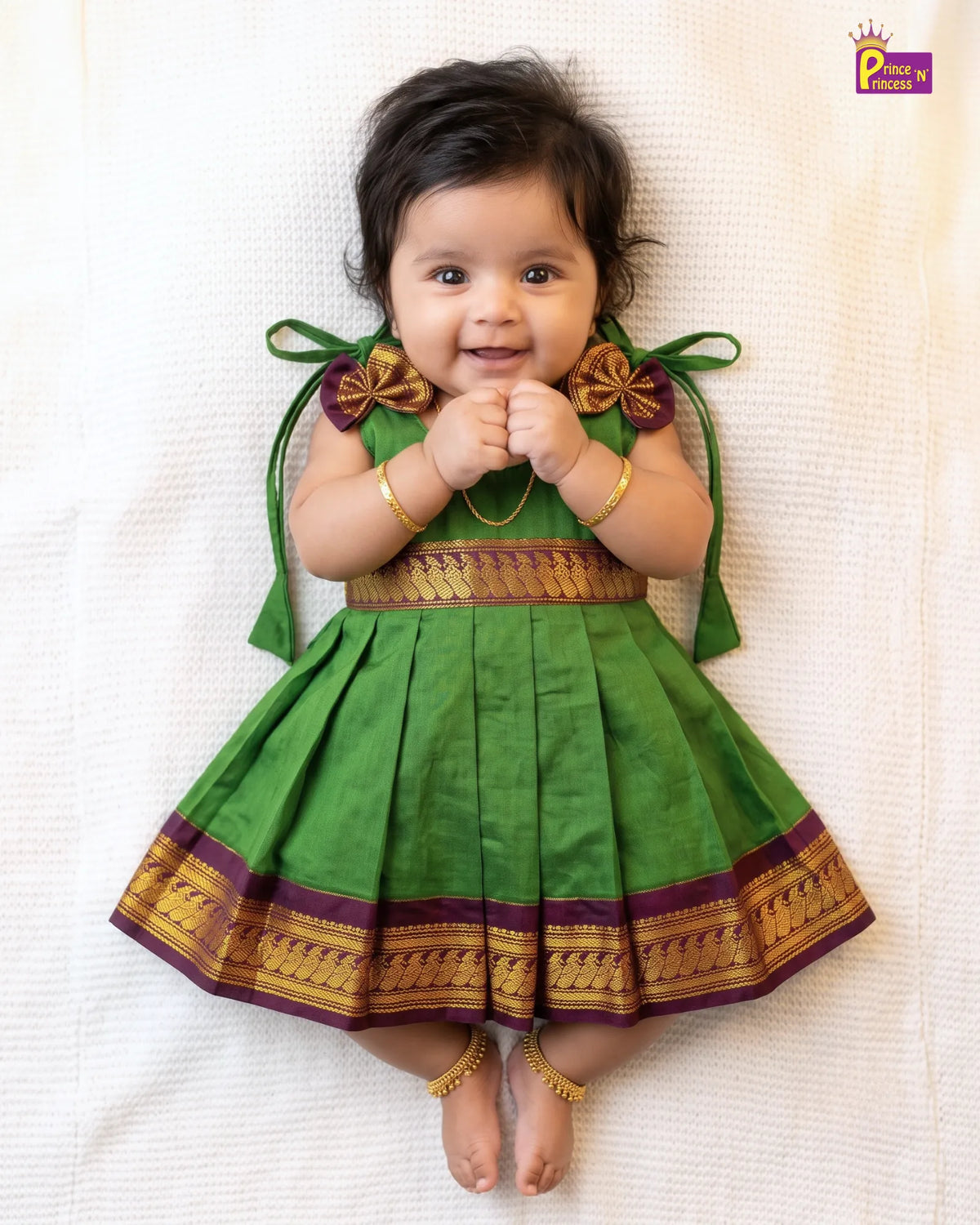 A baby girl lying on a white textured blanket, wearing a green traditional Indian dress with gold and purple borders, bow details on the shoulders, and gold ankle bracelets.