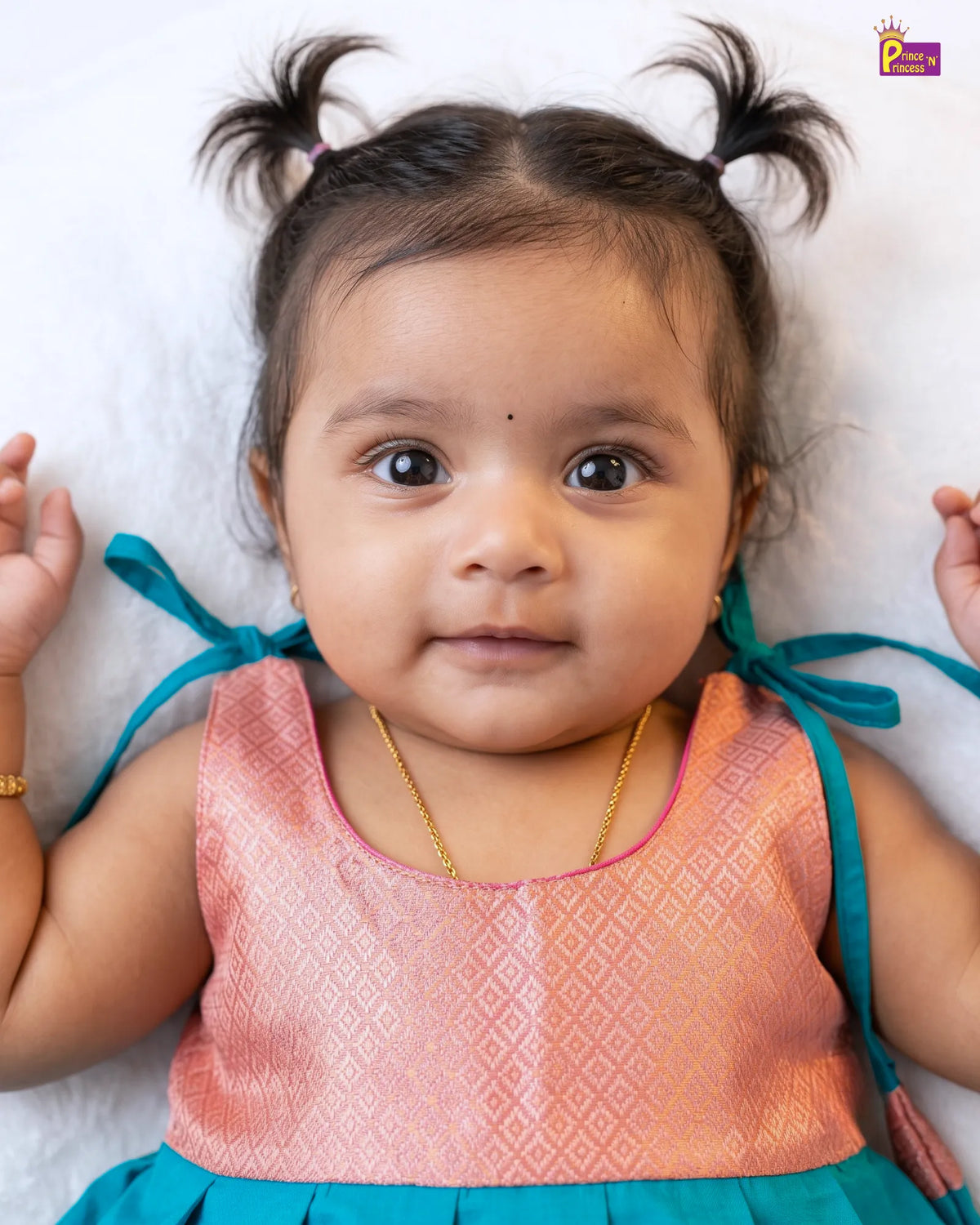 A baby girl lying down wearing a traditional Indian outfit with a peach-pink patterned top and a teal skirt, styled with a gold chain necklace.