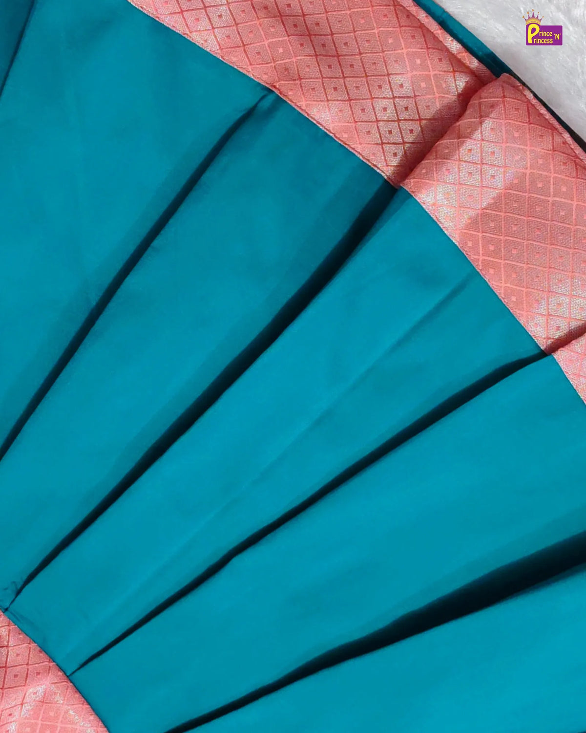 A close-up of a teal blue pleated fabric, likely a saree, with a contrasting pink border featuring a subtle geometric pattern.