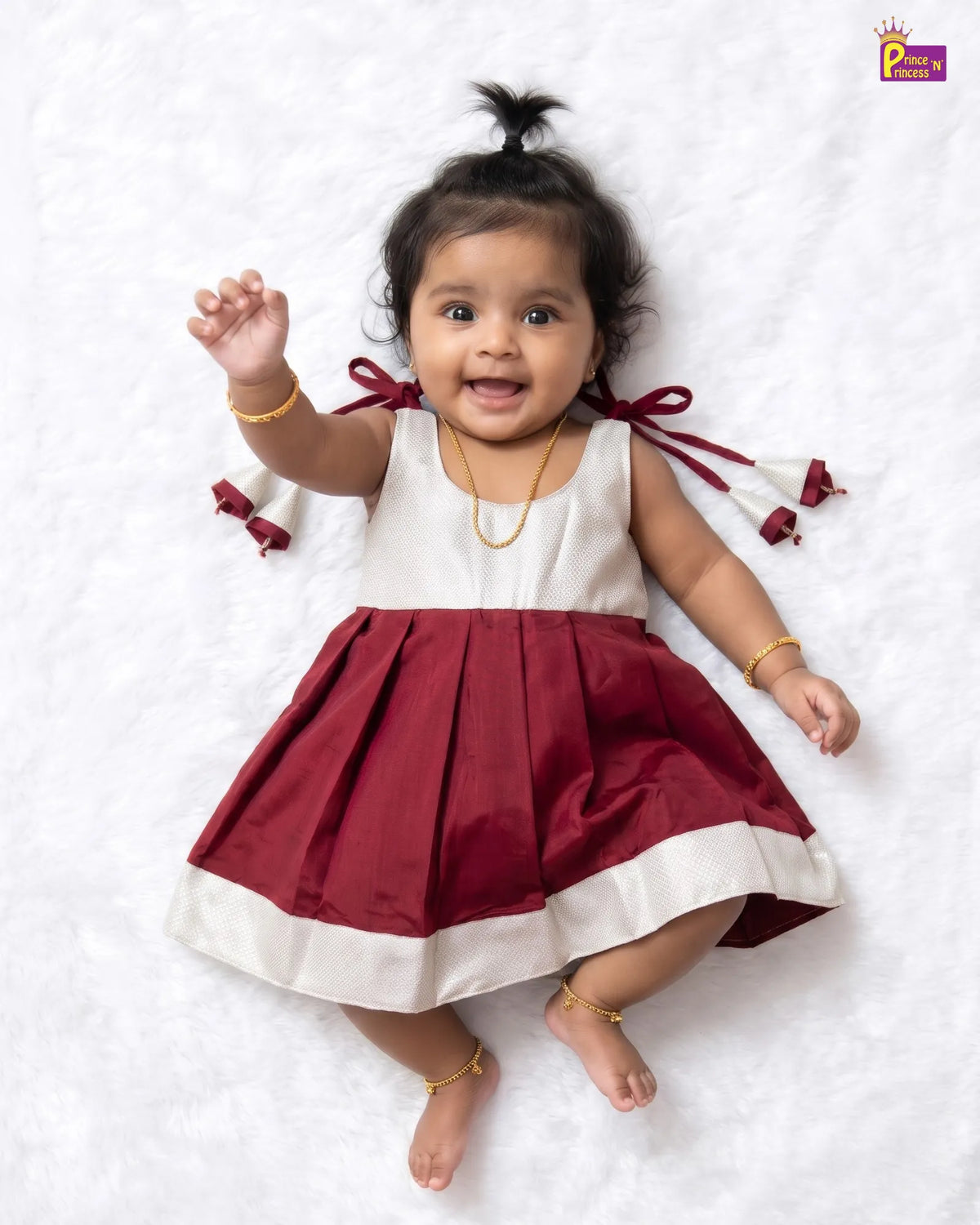 A baby girl lying on a white textured blanket, wearing a sleeveless maroon and white dress with matching maroon ribbon ties on the back. She is accessorized with gold jewelry.