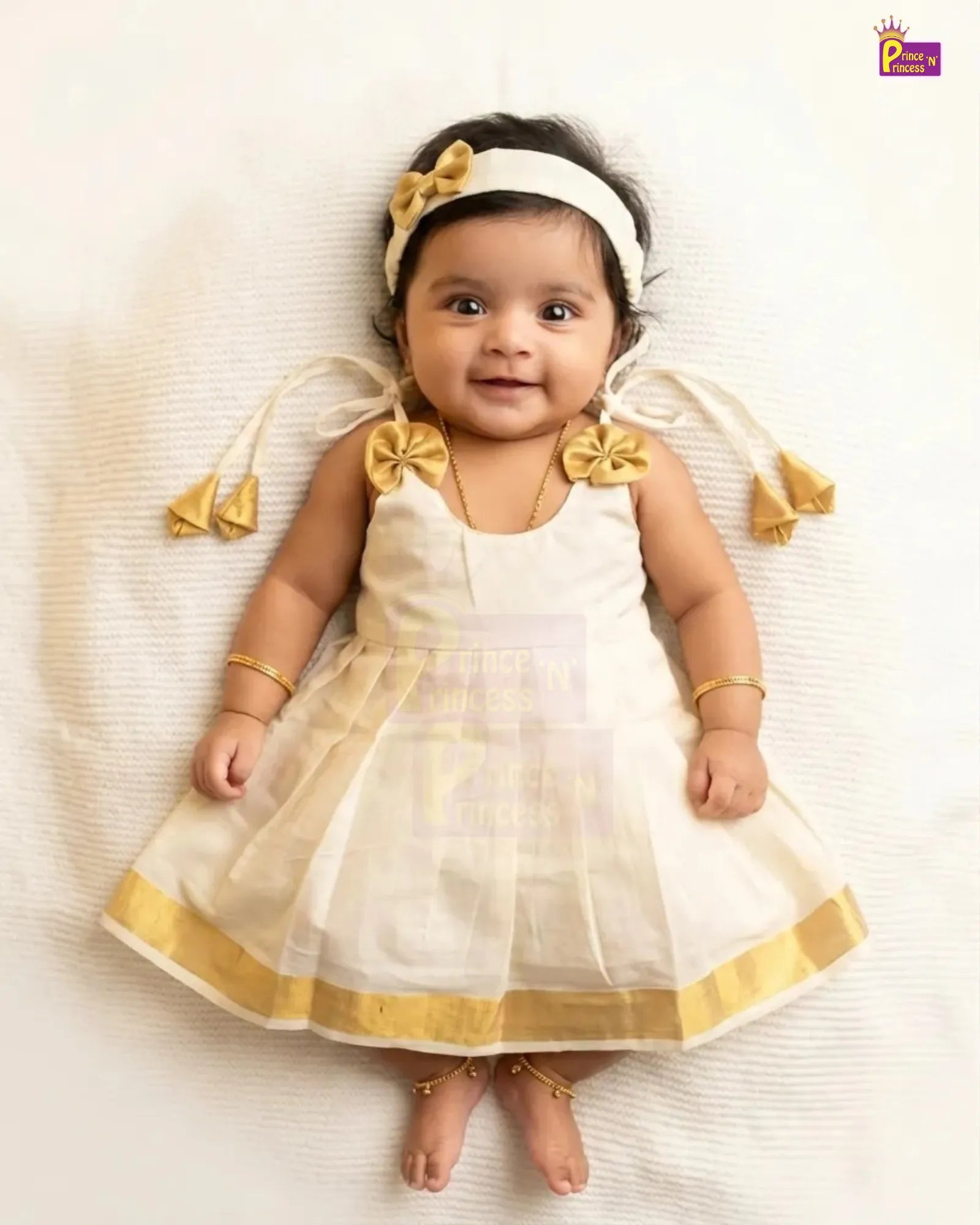 A baby girl lying on her back wearing a white sleeveless dress with gold trim, a white headband with a gold bow, and gold jewelry, set against a white textured background.