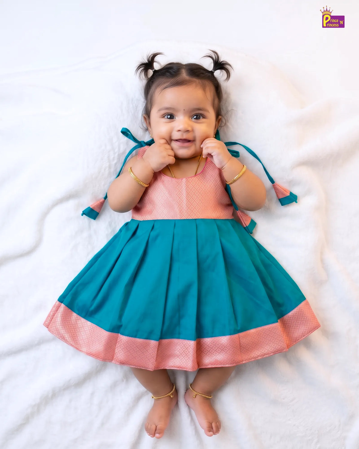 A toddler girl lying on a white blanket wearing a two-tone teal and pink traditional dress with gold bangles and anklets.