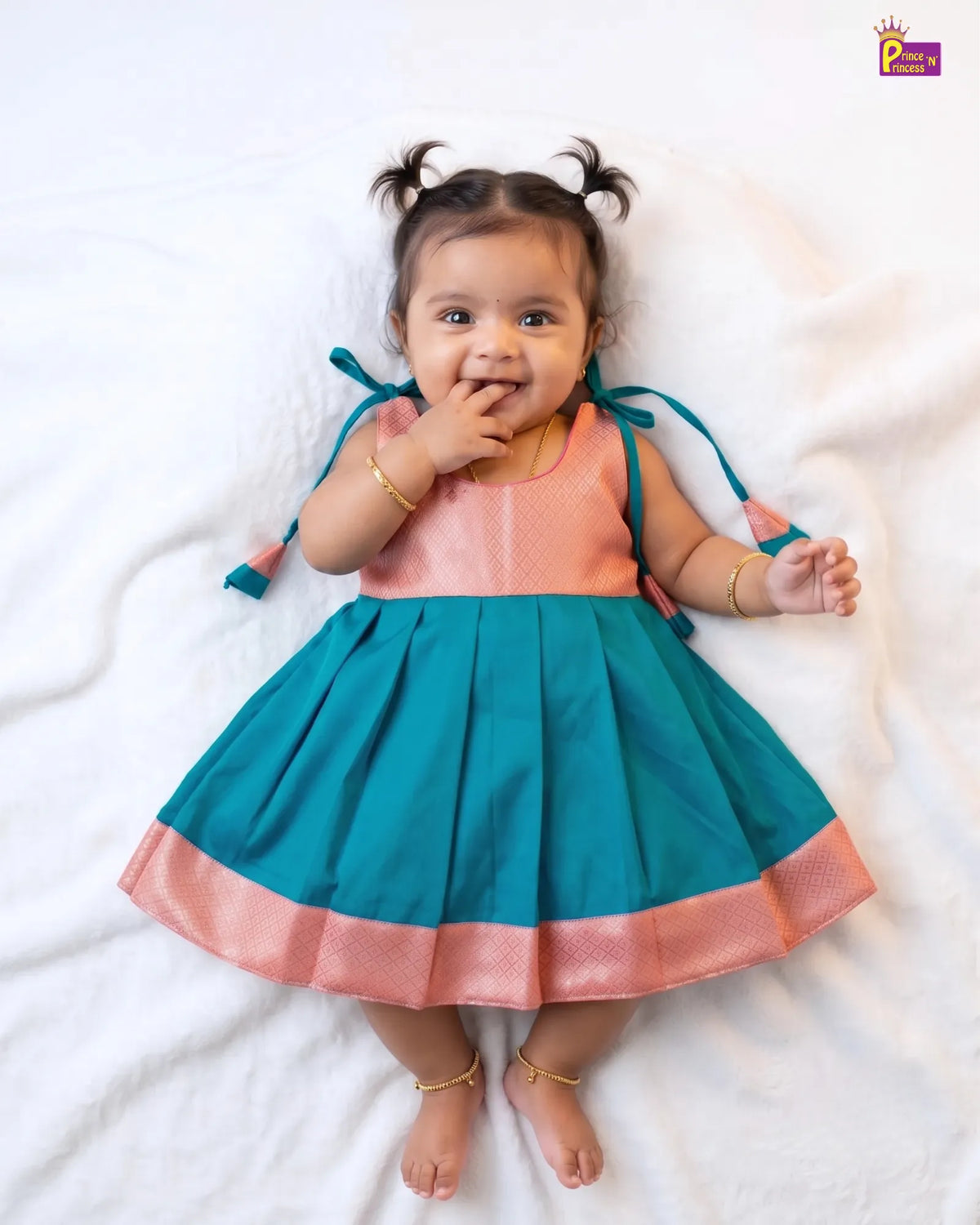 A baby girl lying on a white blanket, wearing a traditional Indian dress with a peach-colored bodice and a teal-colored pleated skirt with a peach border. She has gold jewelry and her hair is styled in two small pigtails.