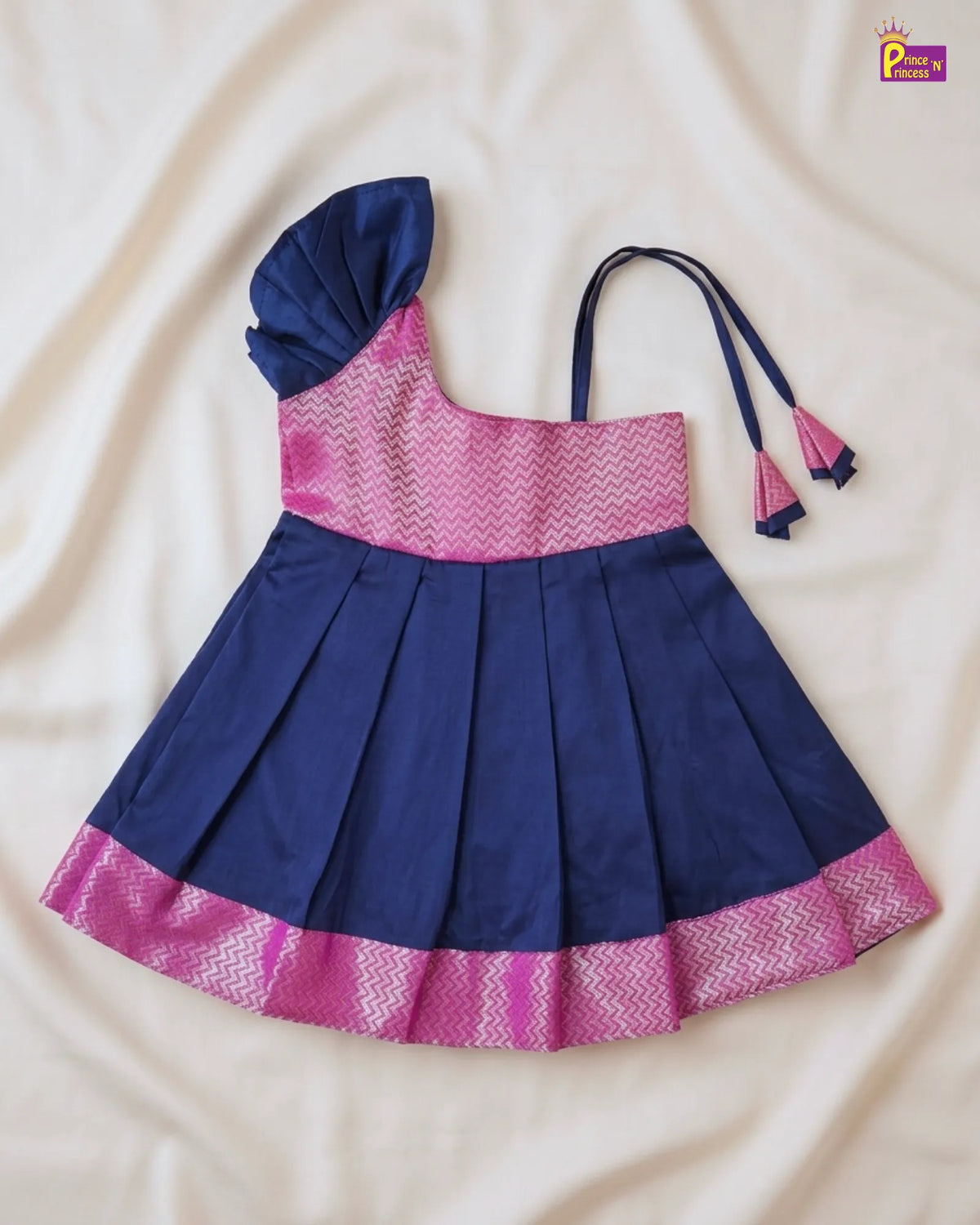 A navy blue and pink traditional Indian girl's dress laid flat on a cream-colored fabric background. The dress features a navy blue pleated skirt with a pink patterned hem, a pink patterned bodice, a navy blue one-shoulder flap, and matching navy tie straps with pink tassels.