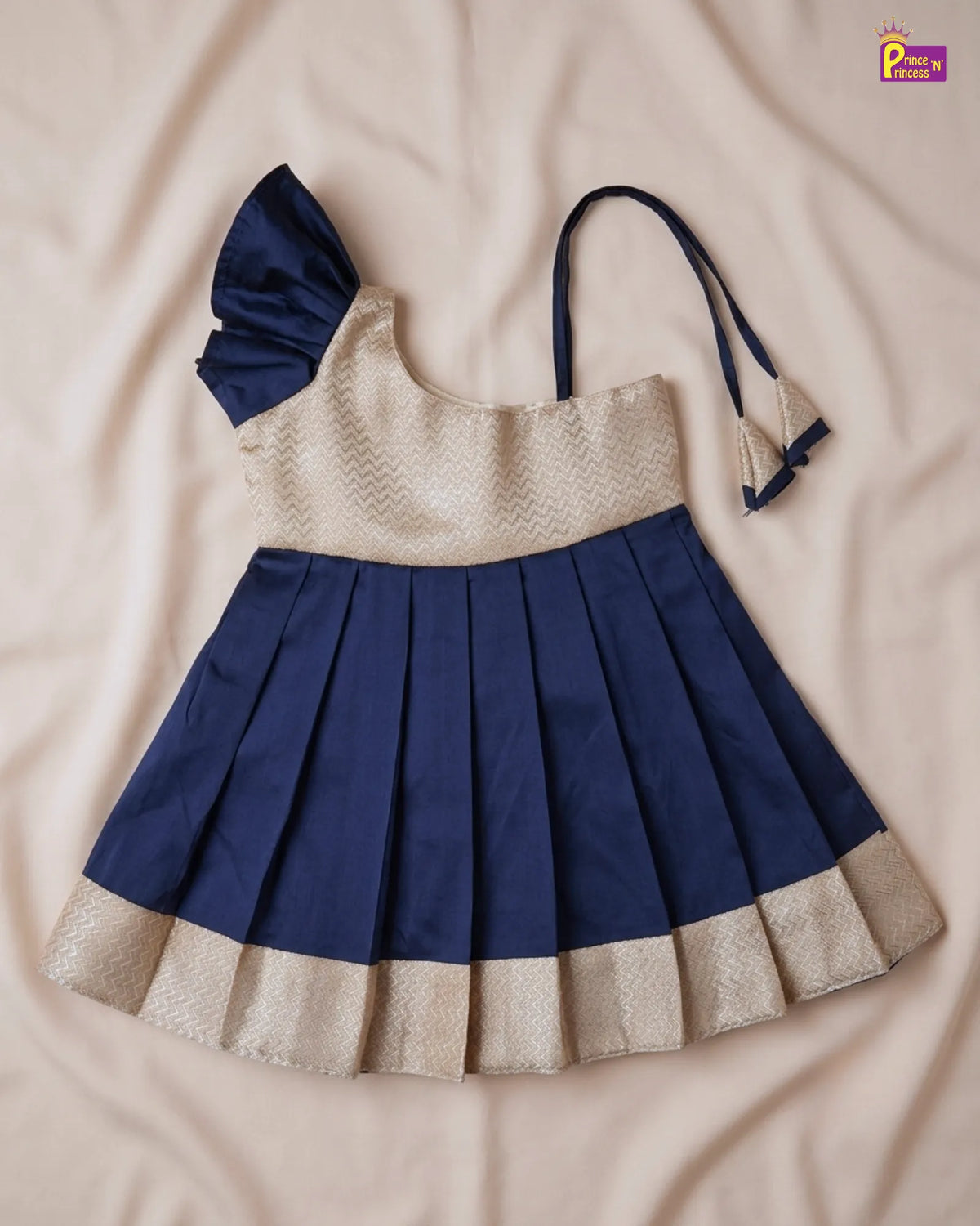 A navy blue and beige toddler girl's dress laid flat on a beige fabric background. The dress features a pleated navy skirt with a beige border, a textured beige bodice, and a navy ruffled shoulder detail with thin shoulder straps.