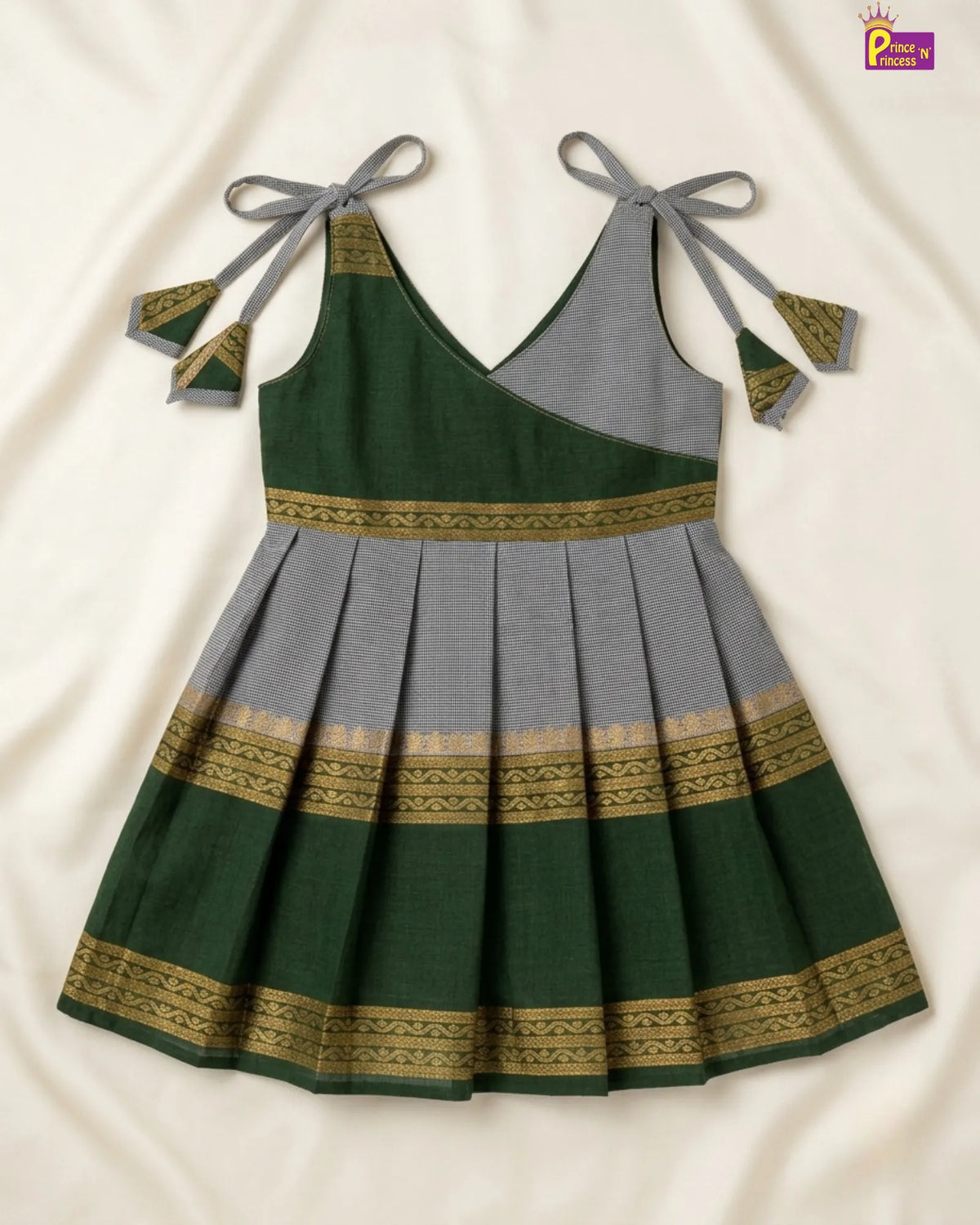A sleeveless pleated dress laid flat on a cream fabric background. The dress has a dark green and gray color-blocked design with gold patterned borders and gray tie straps at the shoulders.