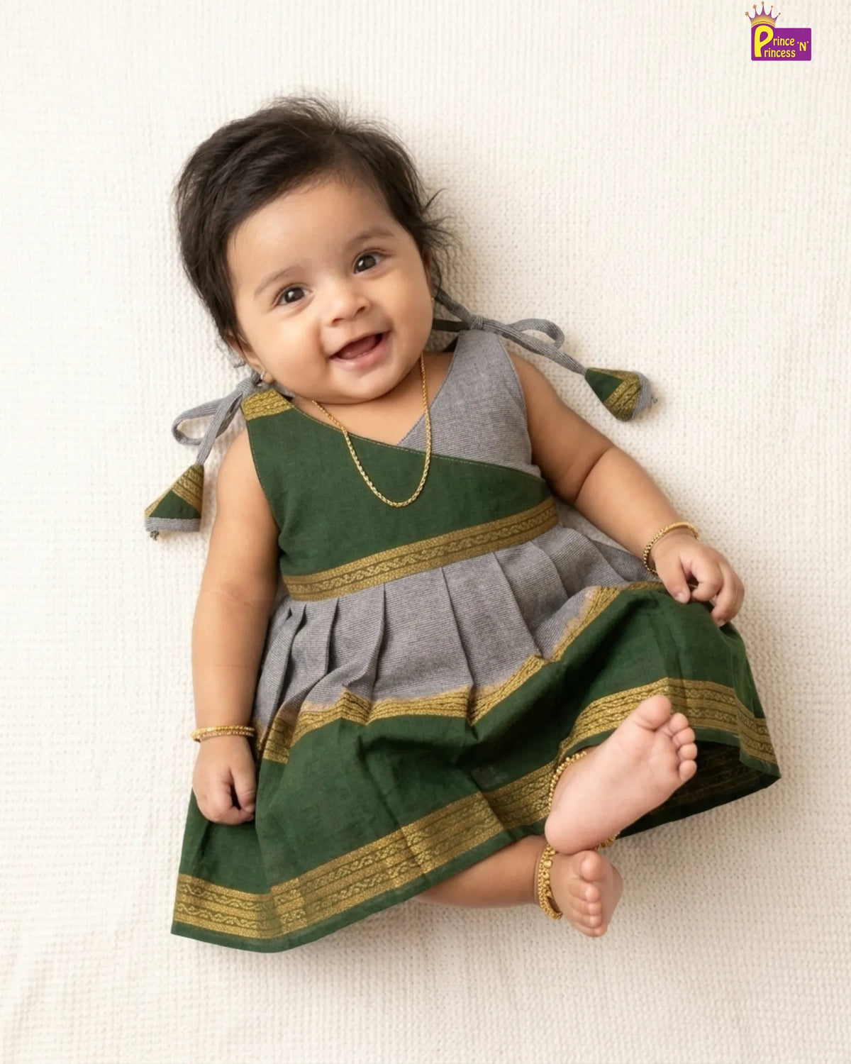 A baby girl lying on a textured cream fabric, wearing a sleeveless traditional-style dress with a green and gray body and gold borders, along with gold jewelry.