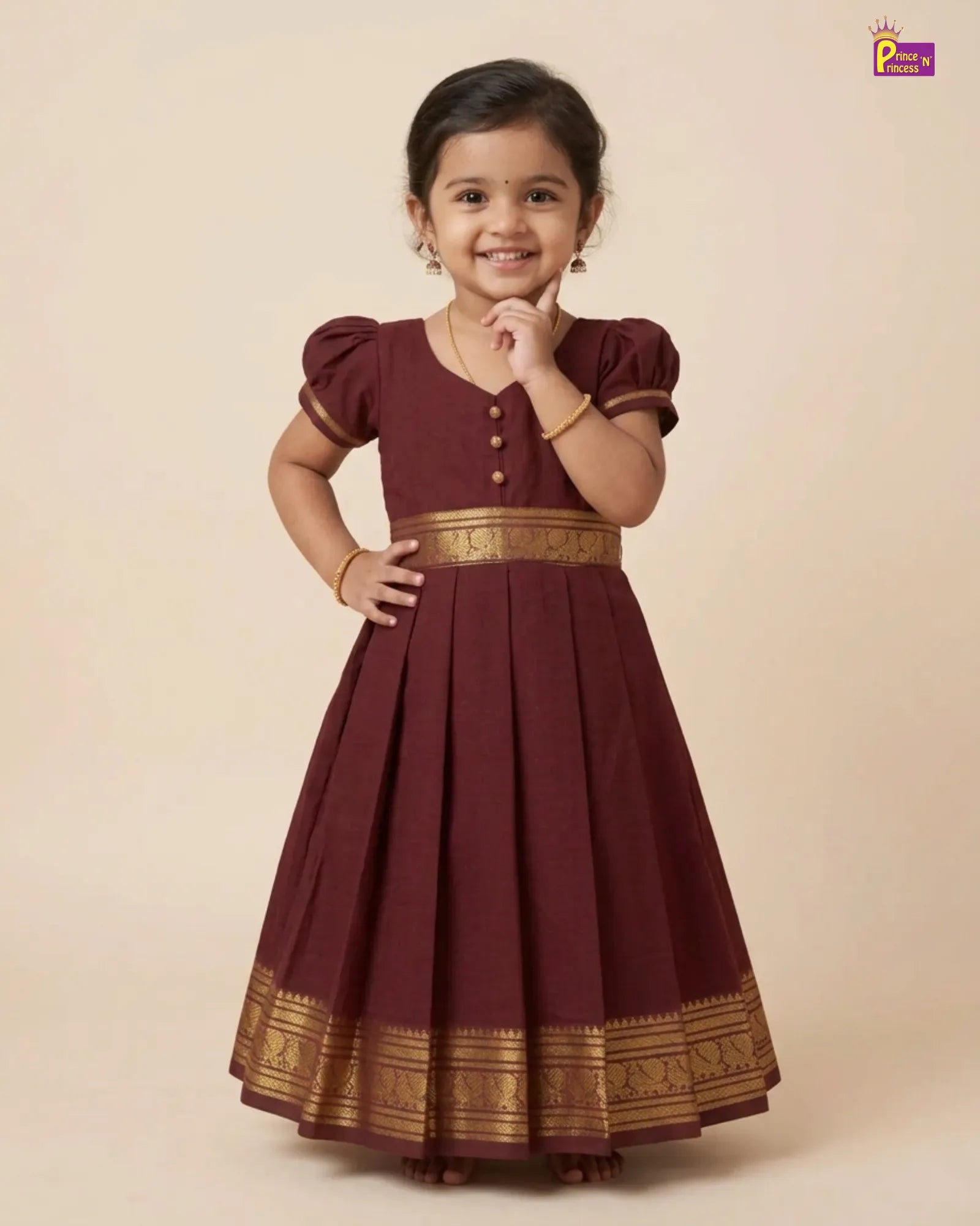 A young girl modeling a maroon A-line ethnic dress with gold zari borders and button details, set against a solid beige studio background.