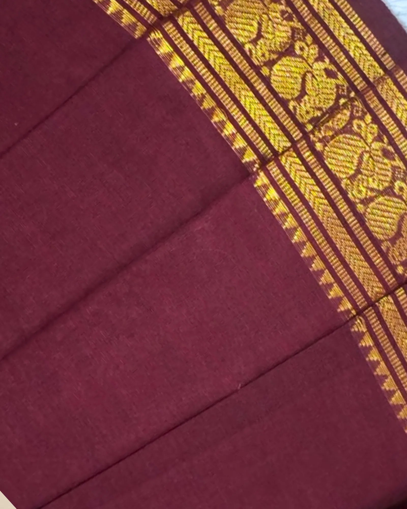 A close-up of a deep maroon fabric with a wide gold woven border featuring traditional floral and geometric patterns.
