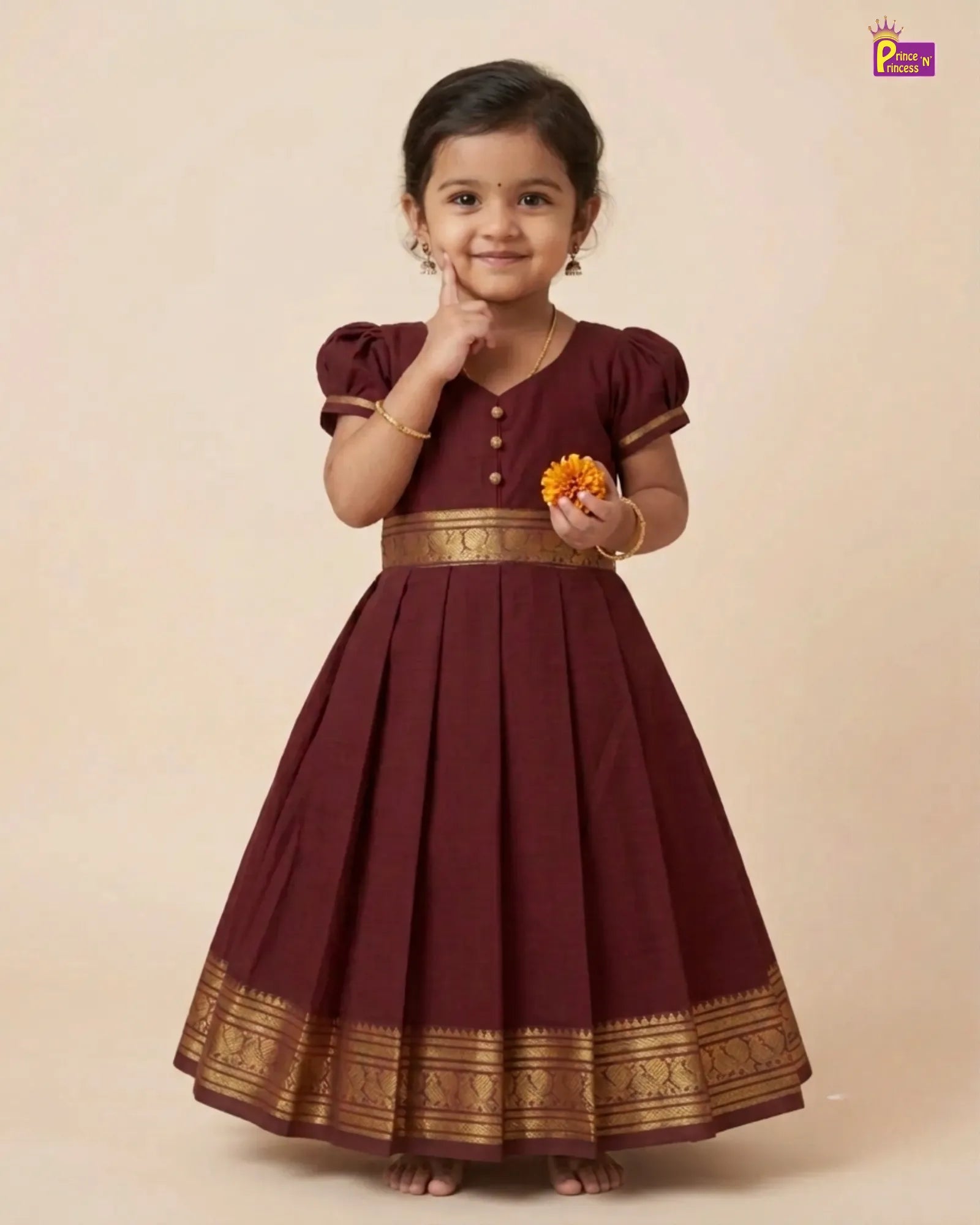 A young girl modeling a traditional maroon A-line dress with gold zari borders and button details, standing against a solid beige studio background.