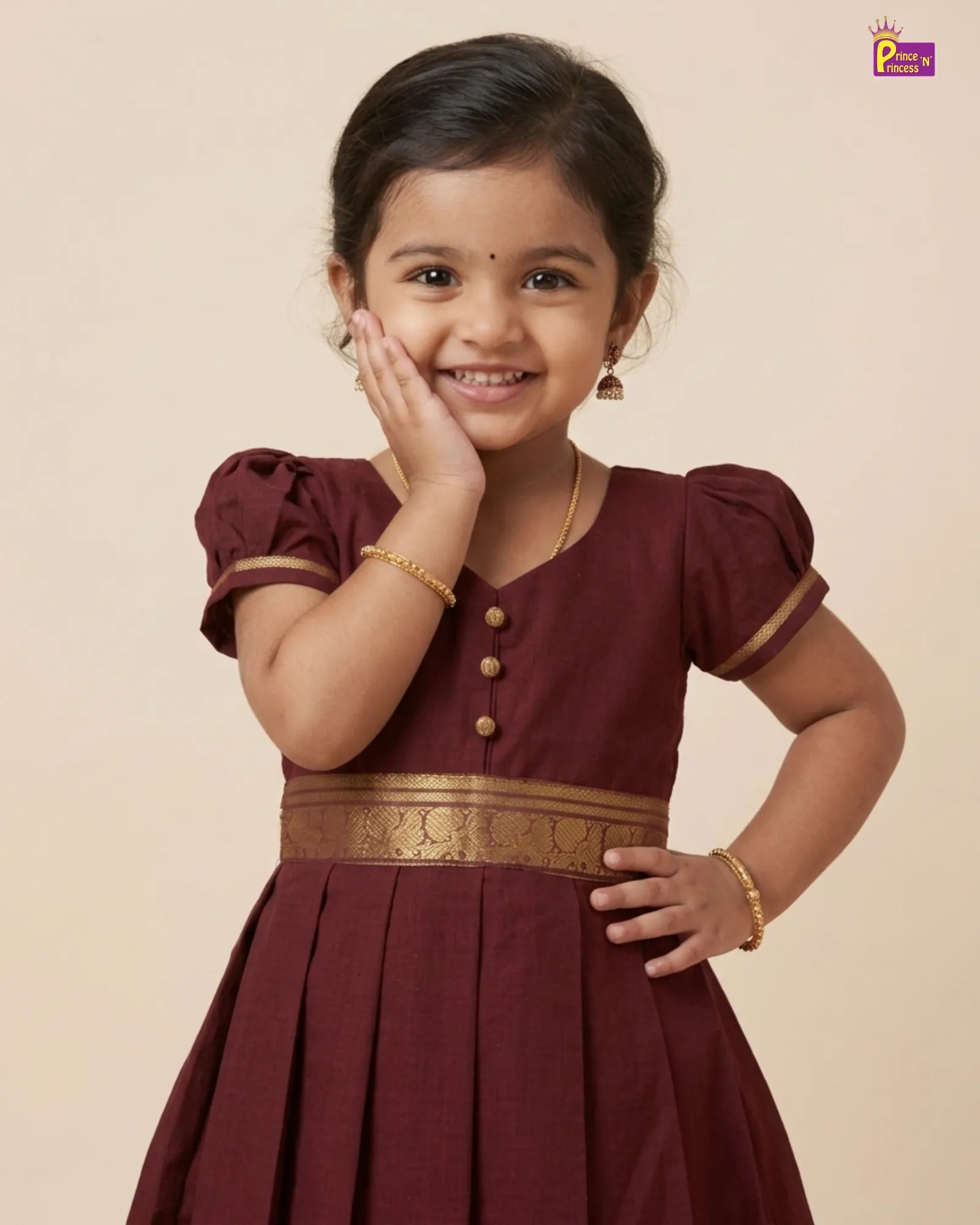 A young girl smiling and posing in a maroon A-line dress with gold trim and buttons, set against a solid beige background.