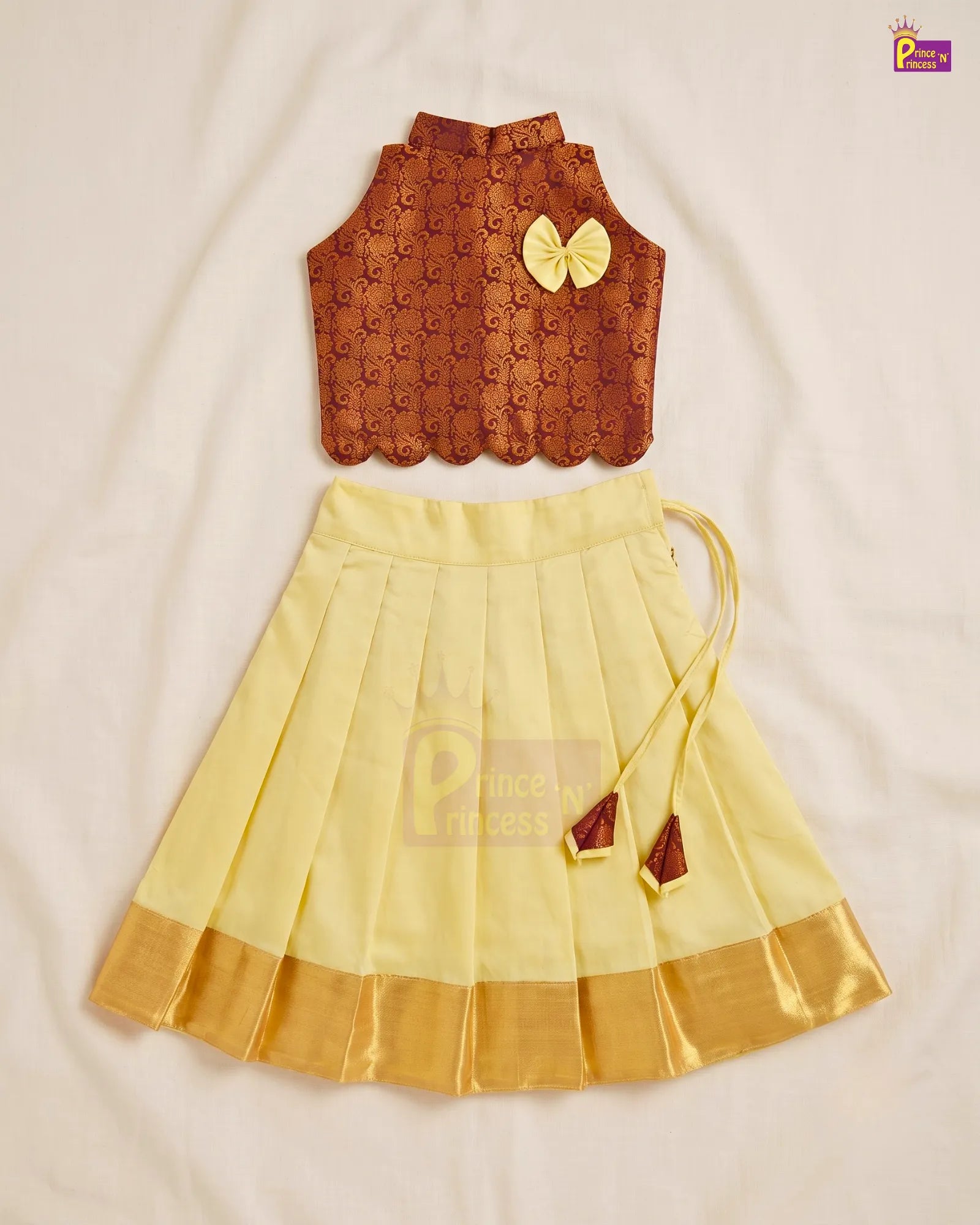 A two-piece children's traditional outfit laid flat on a white fabric background, featuring a brown patterned sleeveless top with a yellow bow accent and a pleated light yellow skirt with a gold zari border and tie details.