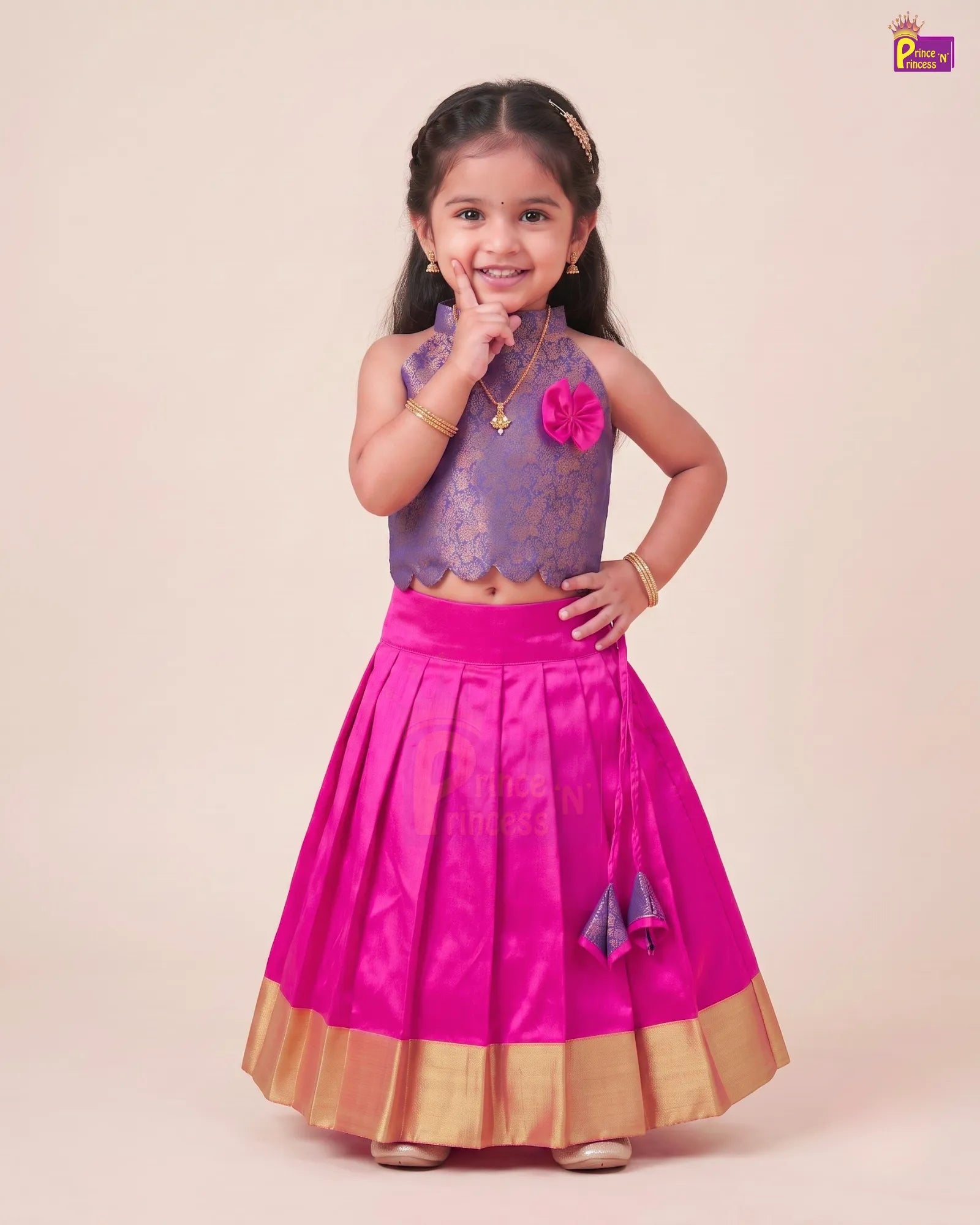 A young girl modeling a traditional Indian ethnic outfit consisting of a purple embroidered sleeveless top and a bright pink pleated skirt with gold zari borders, set against a solid light beige background.