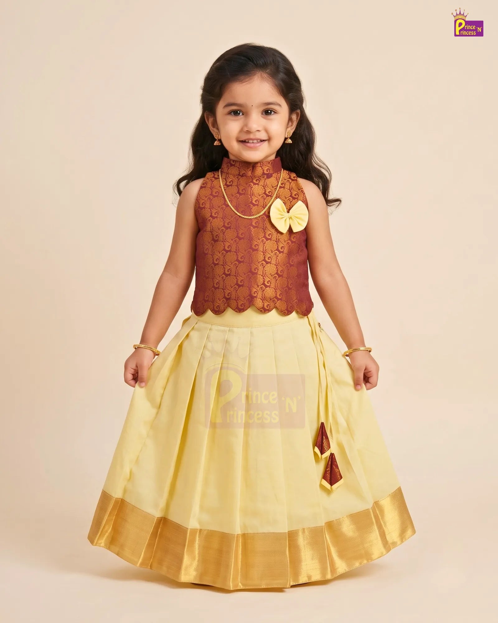 A young girl modeling a traditional Indian ethnic dress consisting of a maroon embroidered sleeveless top and a pleated light yellow skirt with a gold border, set against a solid beige background.