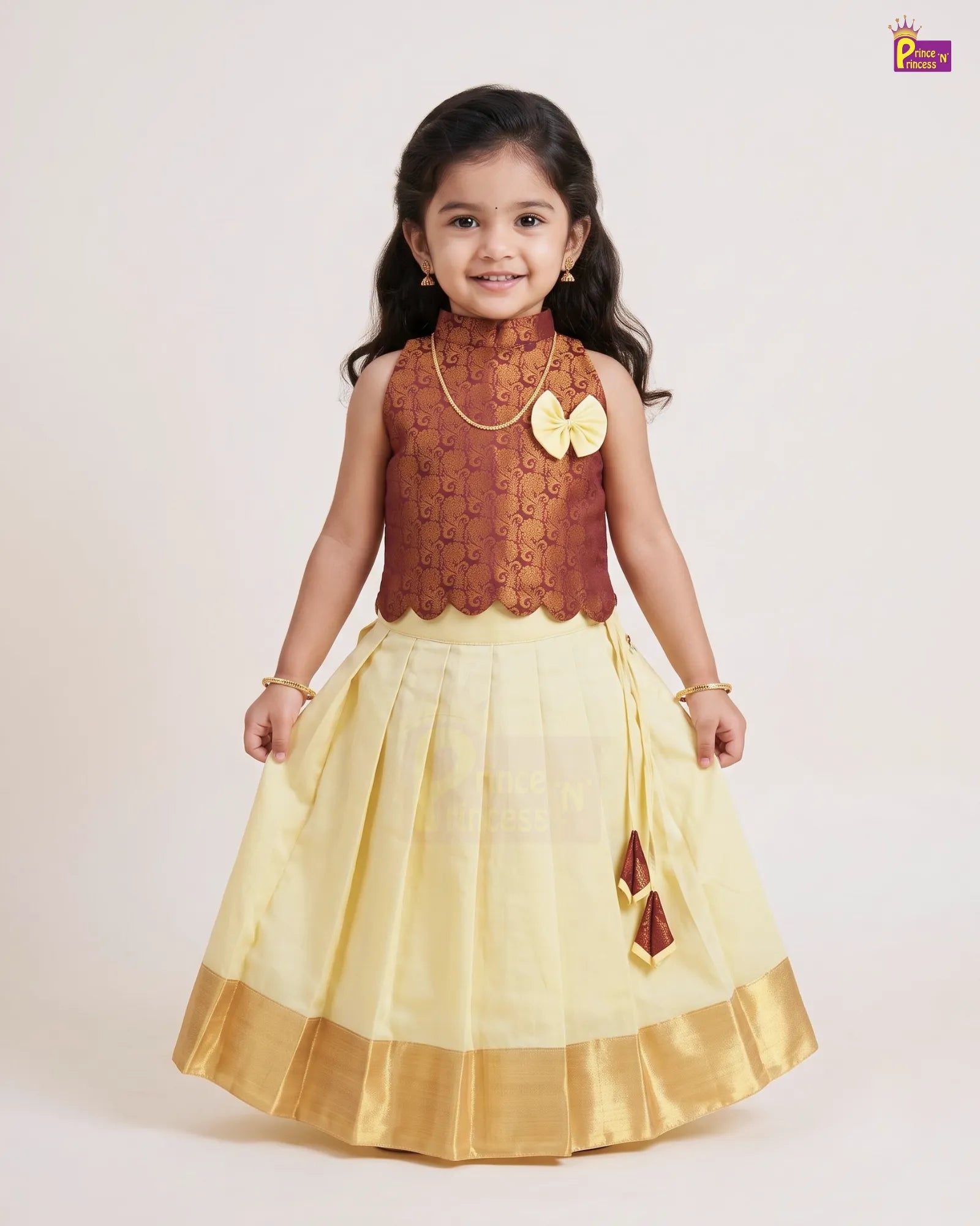 A young girl modeling a traditional Indian outfit consisting of a brown patterned sleeveless top and a pleated light yellow skirt with a gold border, set against a plain light background.