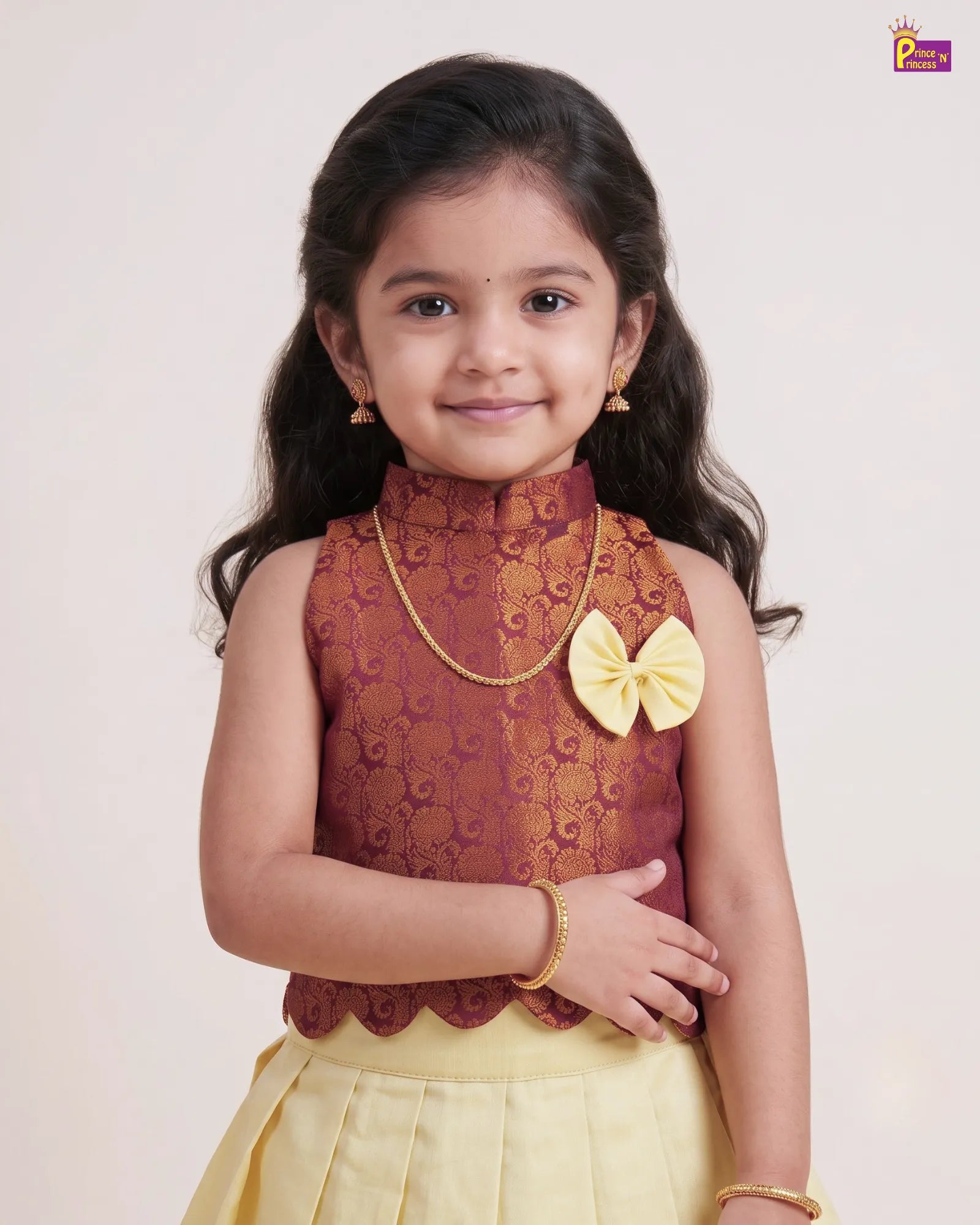 A young girl modeling a traditional Indian outfit featuring a sleeveless maroon top with gold floral embroidery, a yellow bow accent, and a yellow skirt, set against a plain light background.
