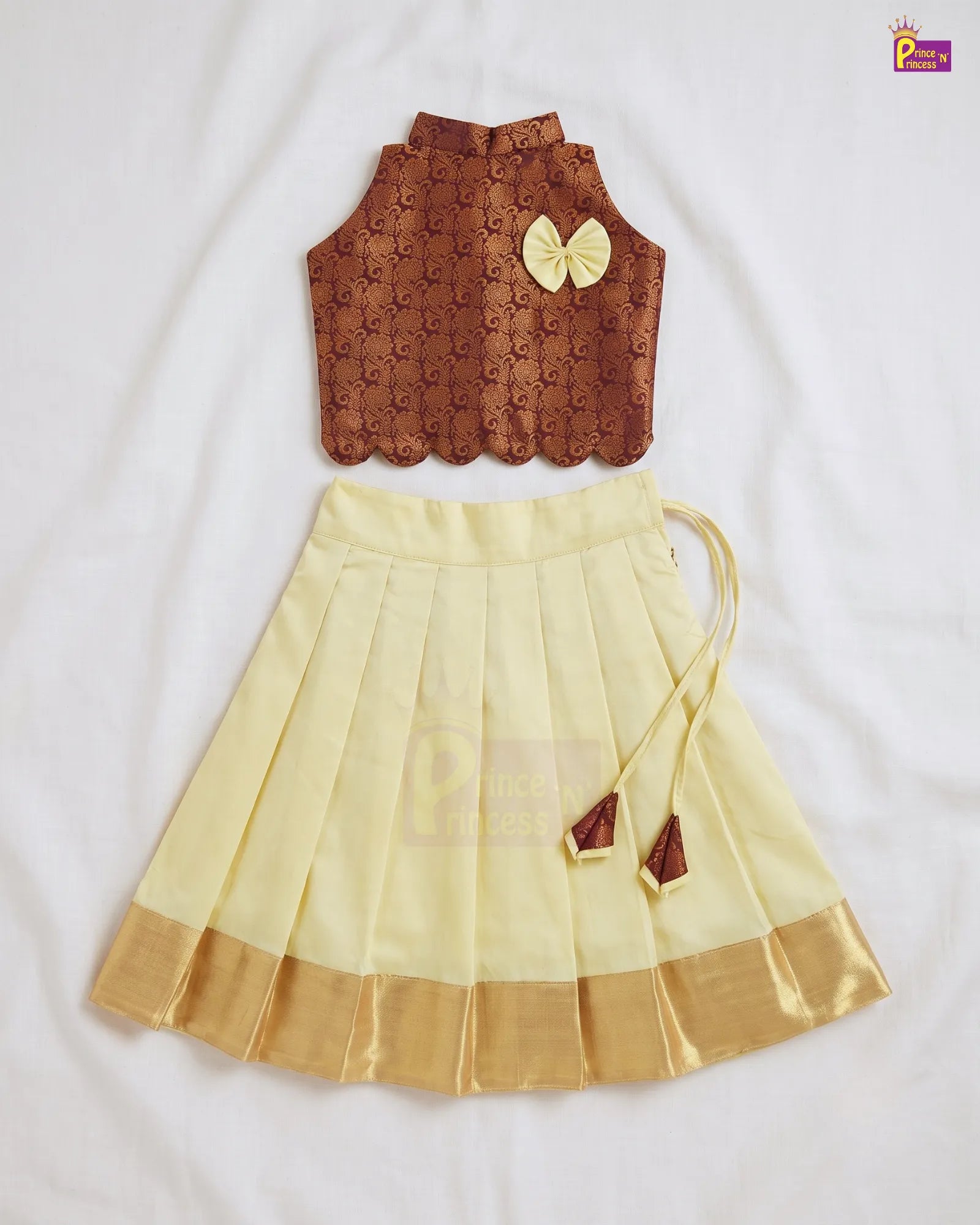 A two-piece children's traditional outfit laid flat on a white fabric background. The top is a sleeveless brown embroidered vest with a scalloped hem and a yellow bow accent.