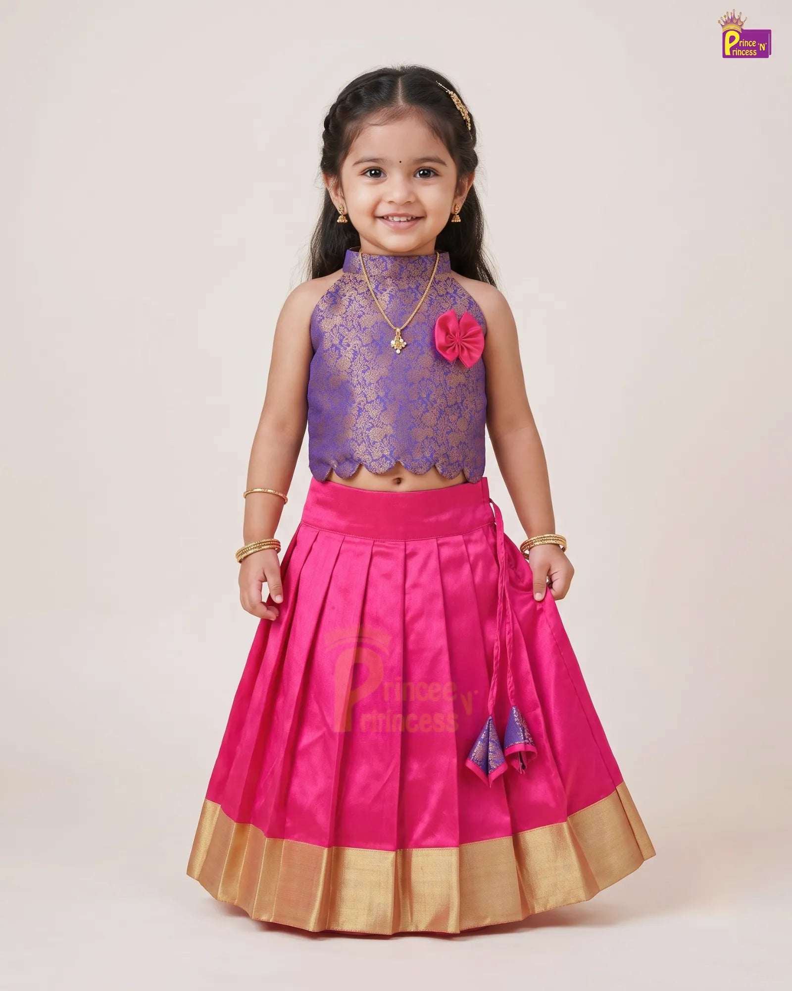 A young girl modeling a traditional Indian outfit consisting of a purple lace sleeveless crop top and a bright pink pleated skirt with gold borders, set against a plain beige studio background.