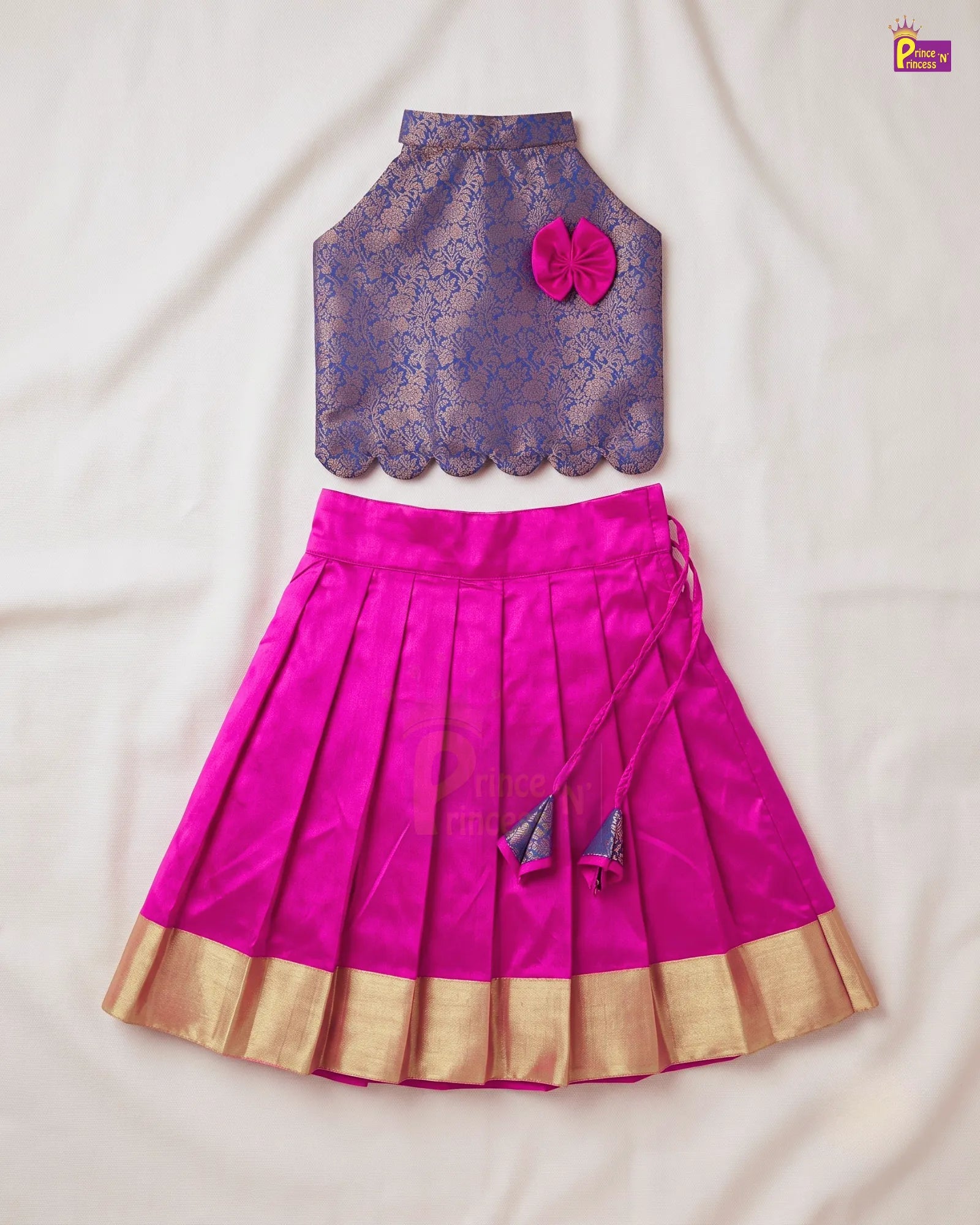 A two-piece children's traditional outfit laid flat on a white fabric background, featuring a purple patterned sleeveless top with a pink flower accent and a bright pink pleated skirt with gold zari borders and tassel ties.