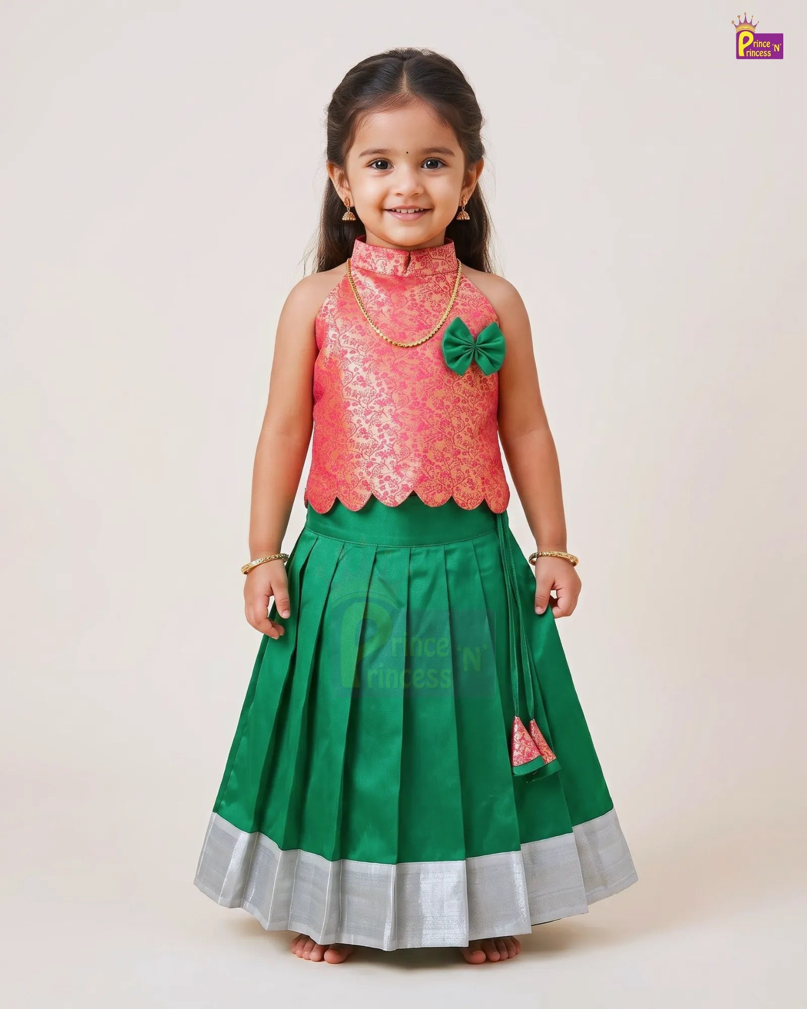 A young girl modeling a traditional Indian ethnic outfit consisting of a pink embroidered sleeveless top and a green pleated skirt with a silver border, set against a plain light beige background.