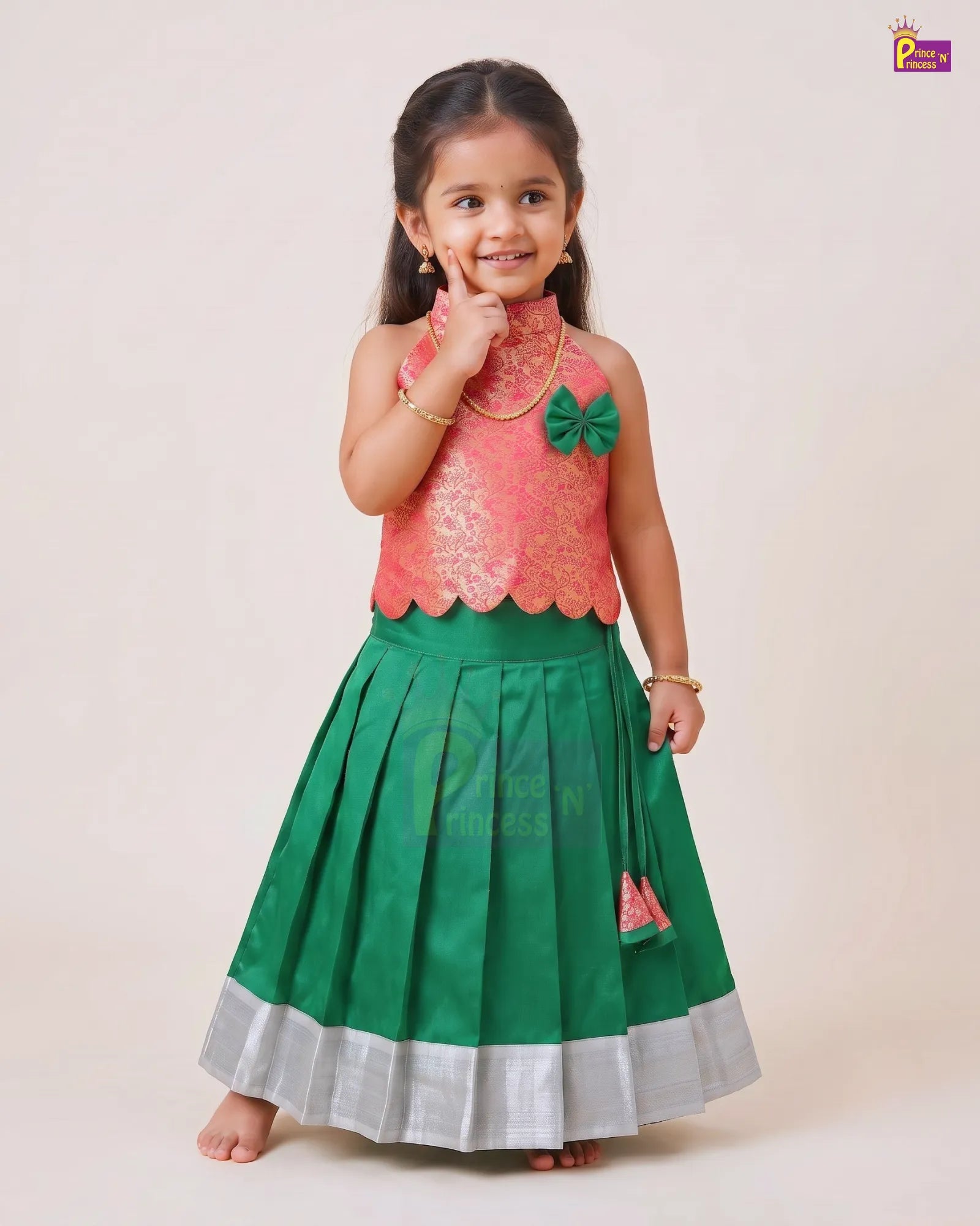 A young girl modeling a traditional Indian ethnic outfit consisting of a coral pink embroidered sleeveless top and a green pleated skirt with a silver border, set against a plain beige studio background.