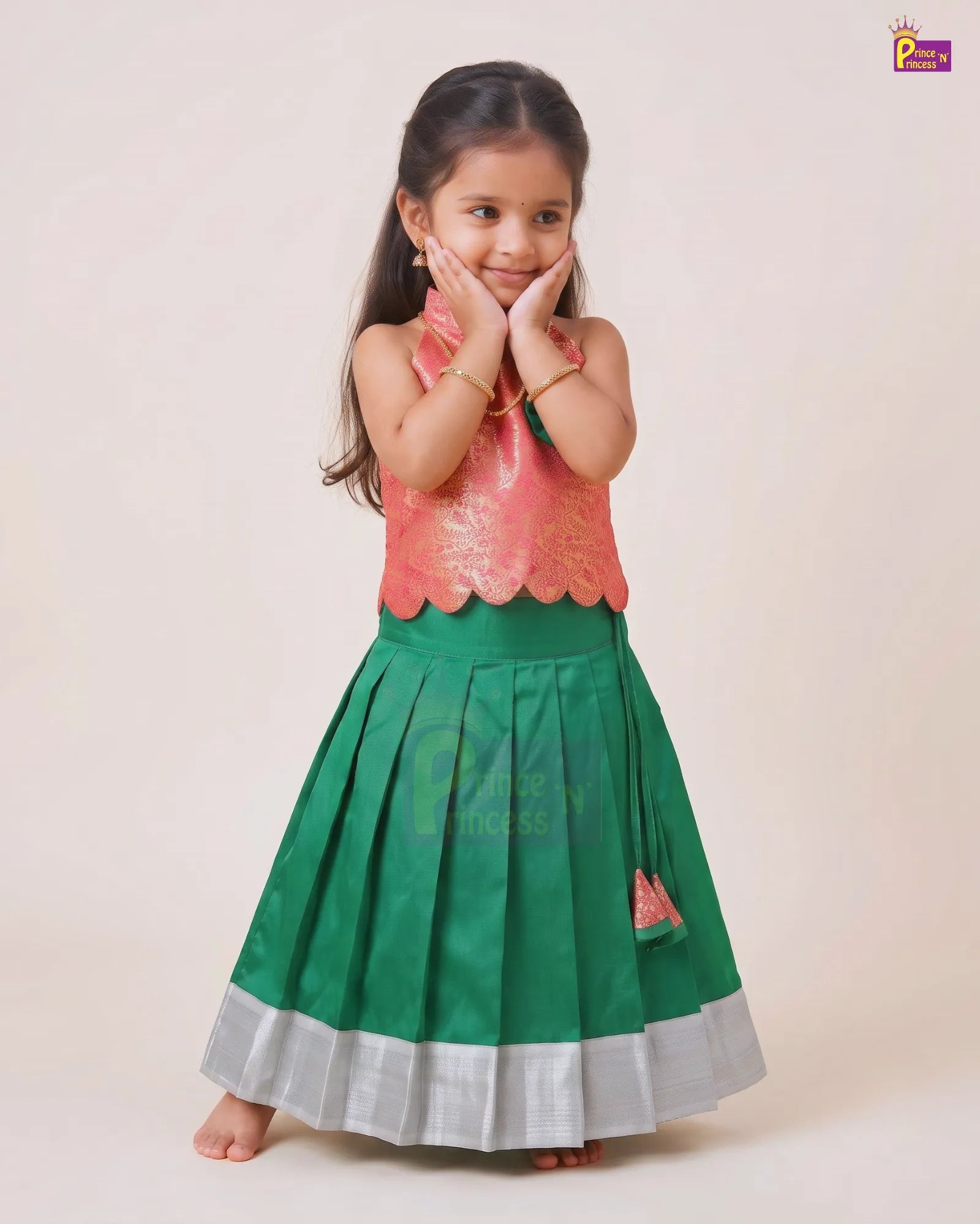 A young girl modeling a traditional Indian ethnic dress with a metallic pink sleeveless top and a pleated green skirt with a silver border, set against a plain beige background.