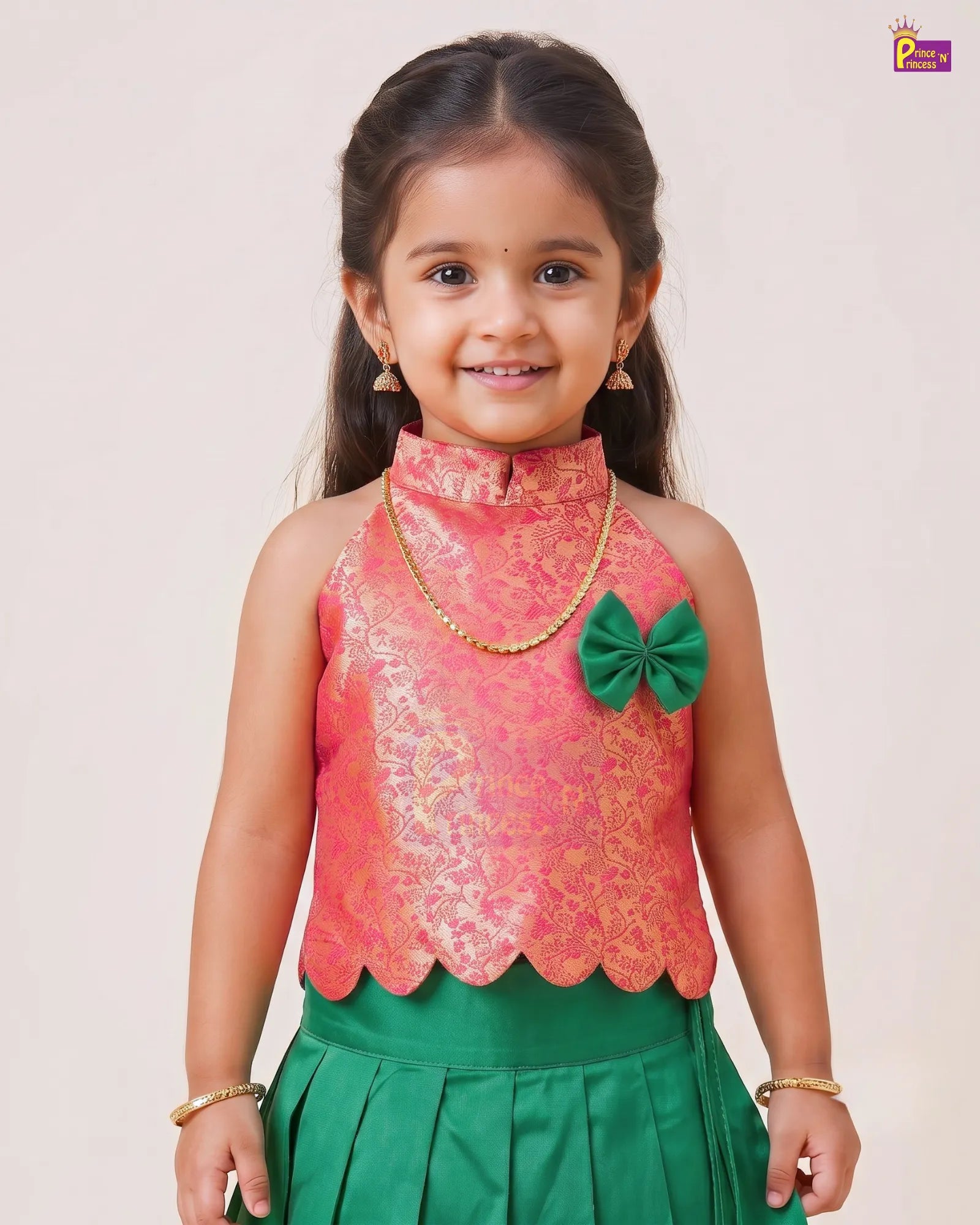 A young girl modeling a traditional Indian outfit consisting of a sleeveless coral pink and gold brocade top with a green bow accent, paired with a green skirt, set against a plain light beige background.