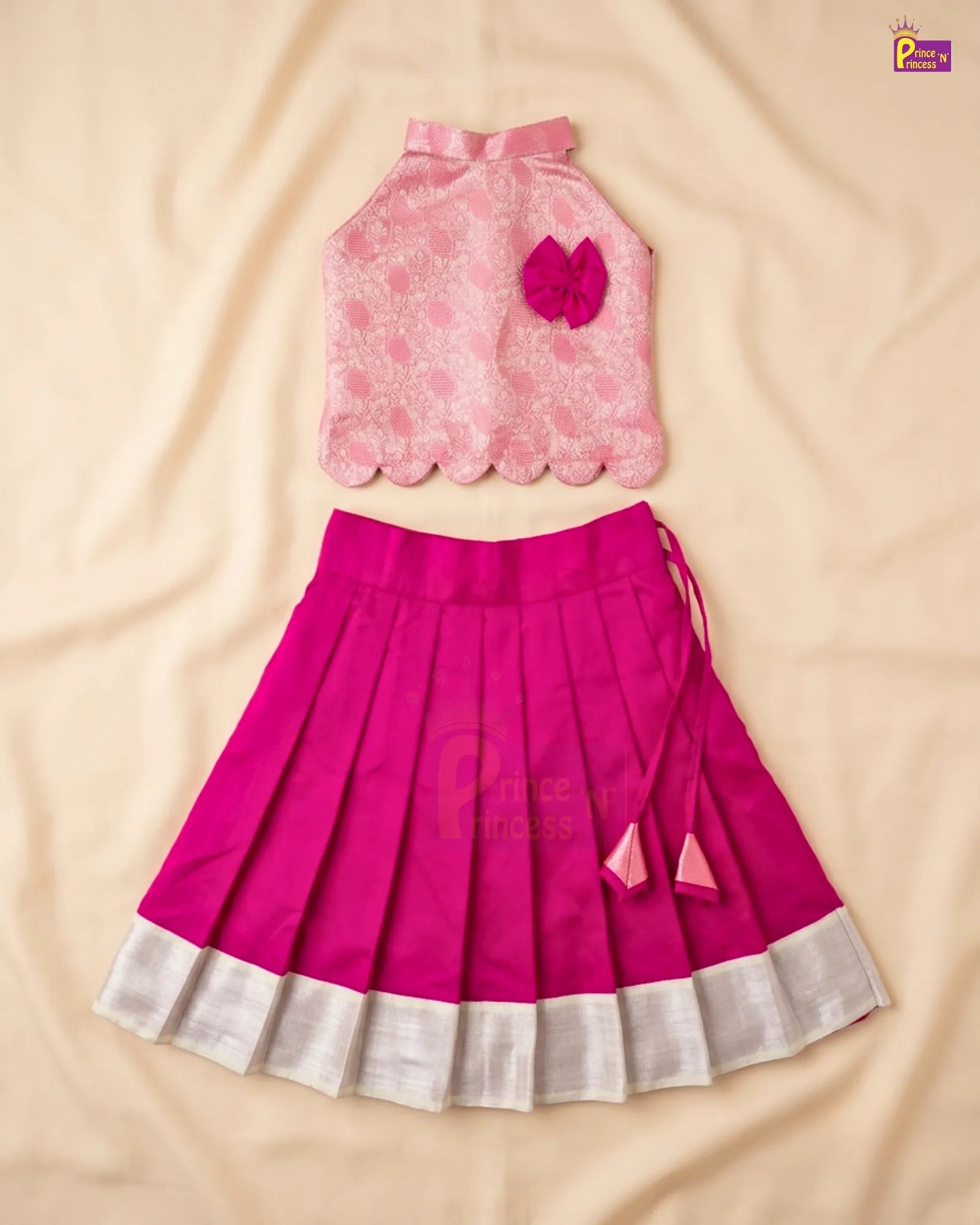 A two-piece girls' traditional outfit laid flat on a beige fabric background, featuring a light pink floral sleeveless top with a scalloped hem and a dark pink pleated skirt with a white border.