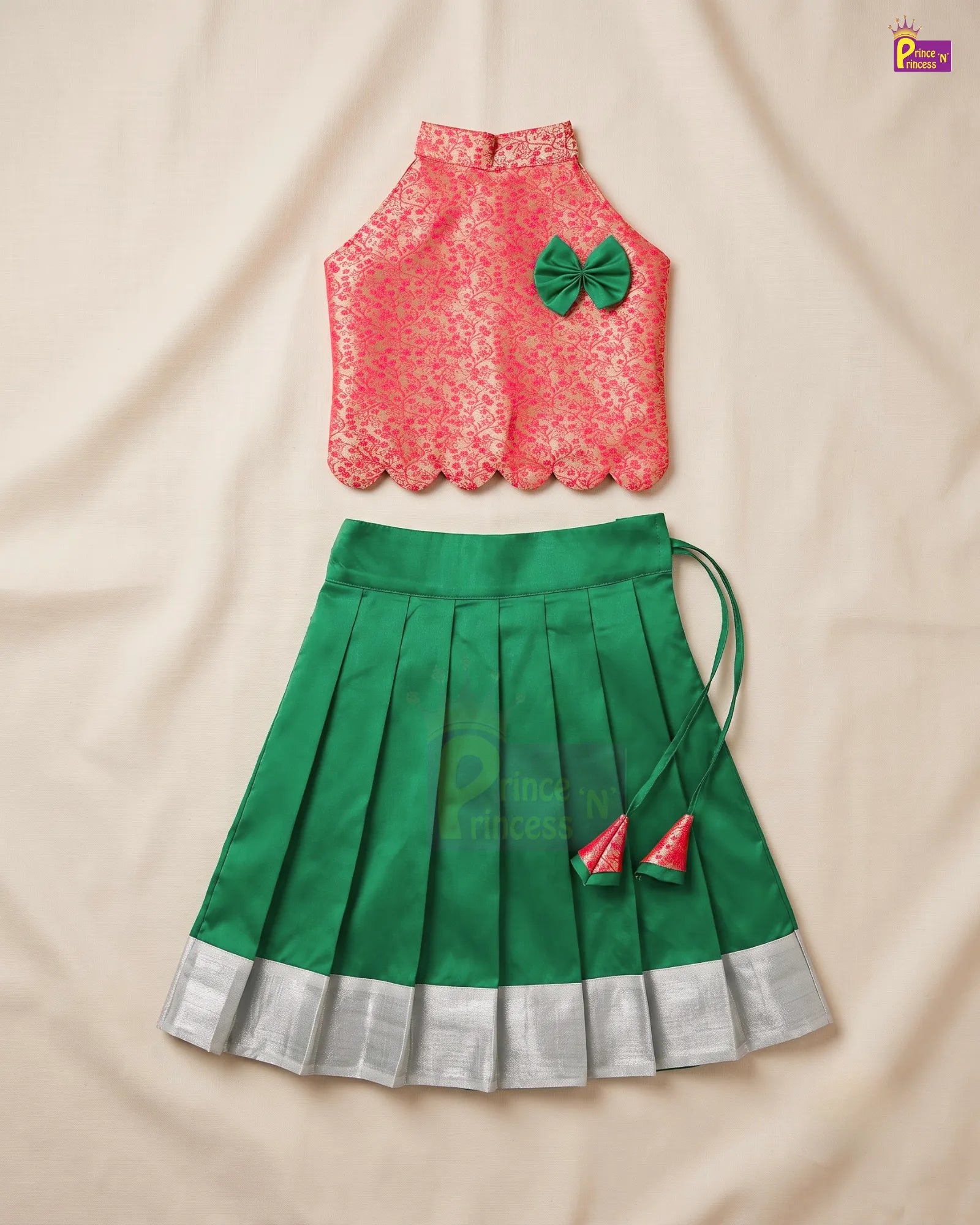 A two-piece girls' traditional outfit laid flat on a beige fabric background. The top is a sleeveless coral-pink lace blouse with scalloped edges and a green bow accent.