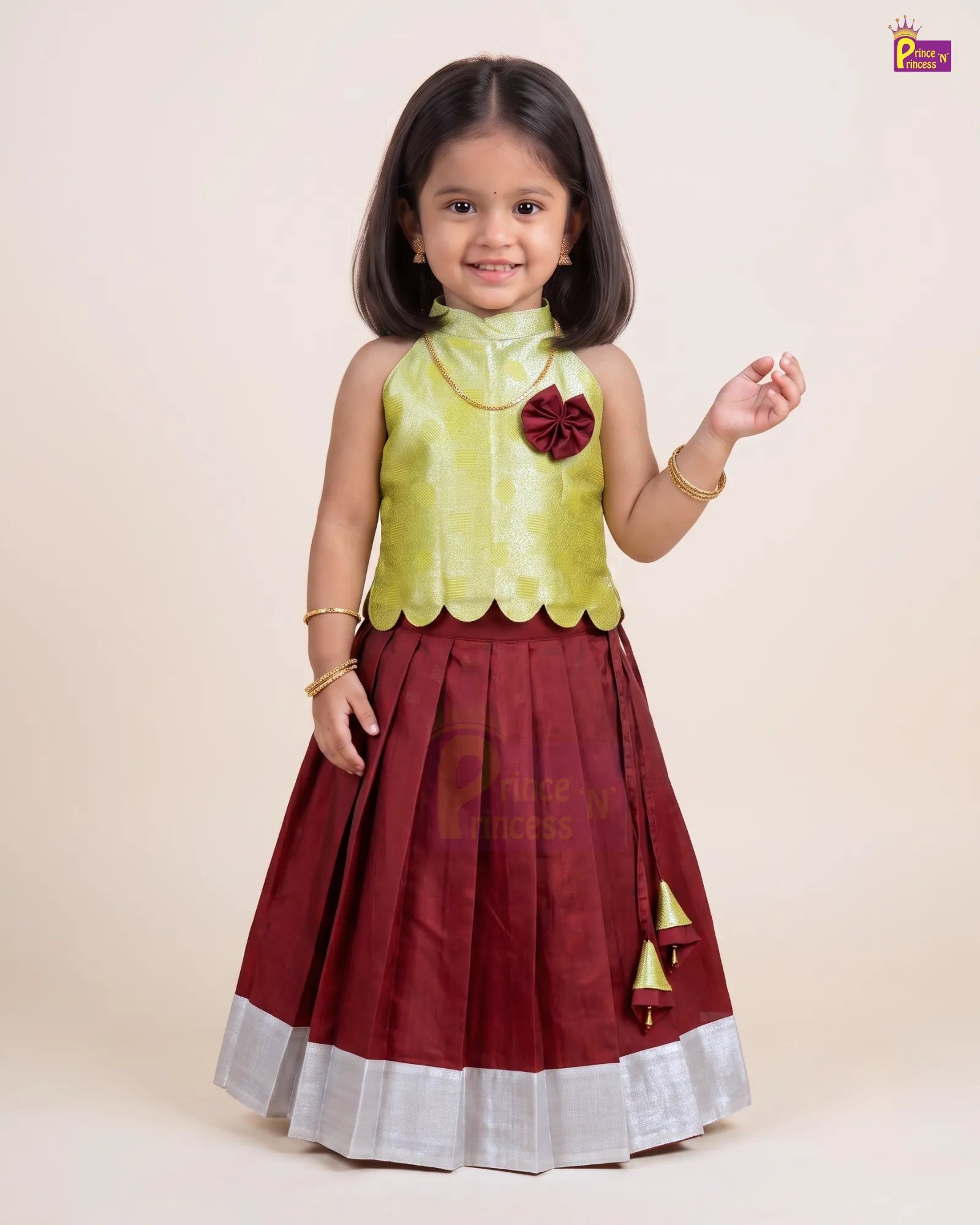 A young girl modeling a traditional Indian dress with a light green sleeveless top and a dark maroon pleated skirt with a silver border and gold tassel details, set against a plain beige background.