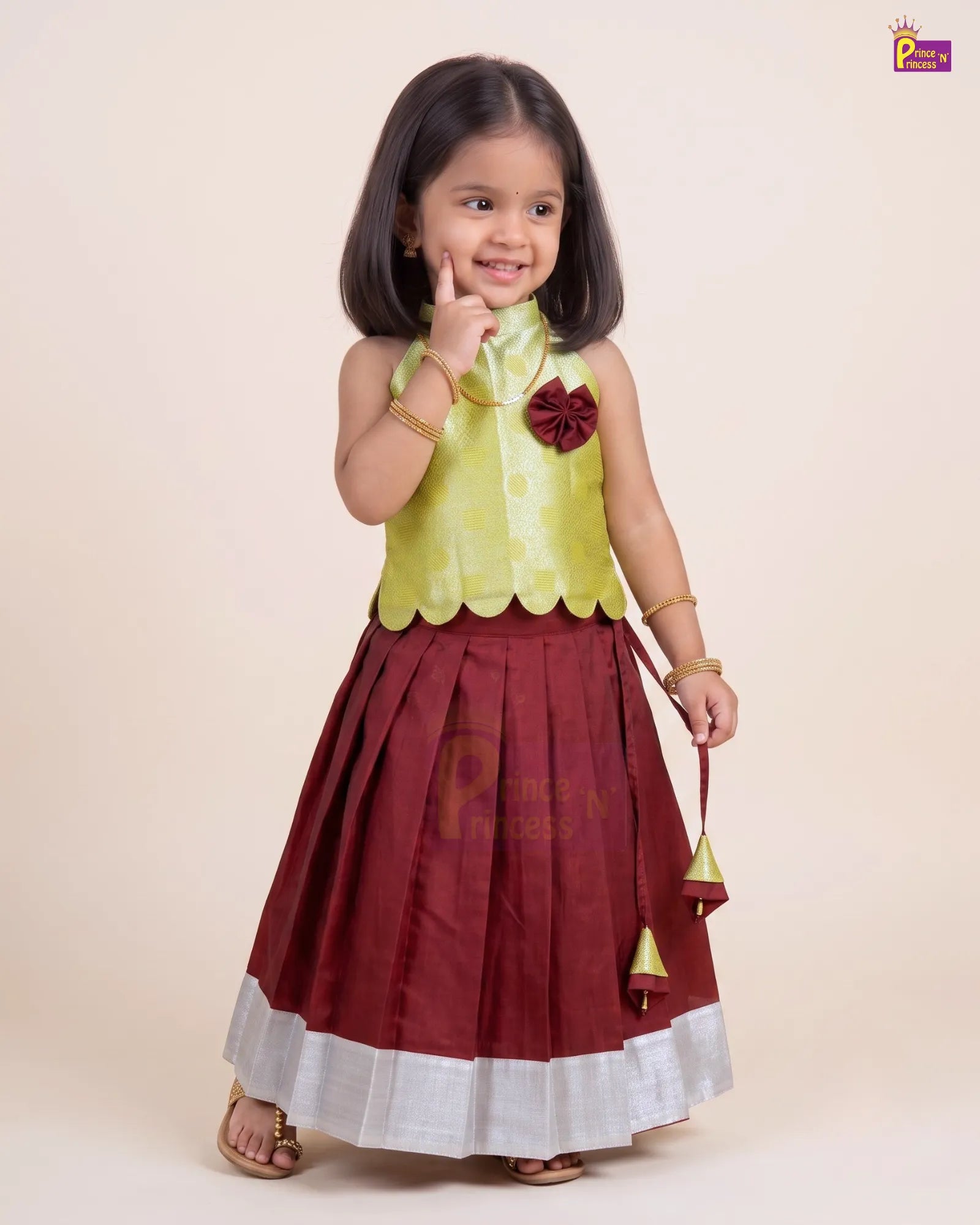 A young girl modeling a traditional Indian outfit consisting of a light yellow-green sleeveless top with a maroon bow accent and a pleated maroon skirt with a white border, holding a small maroon and gold jhumka.