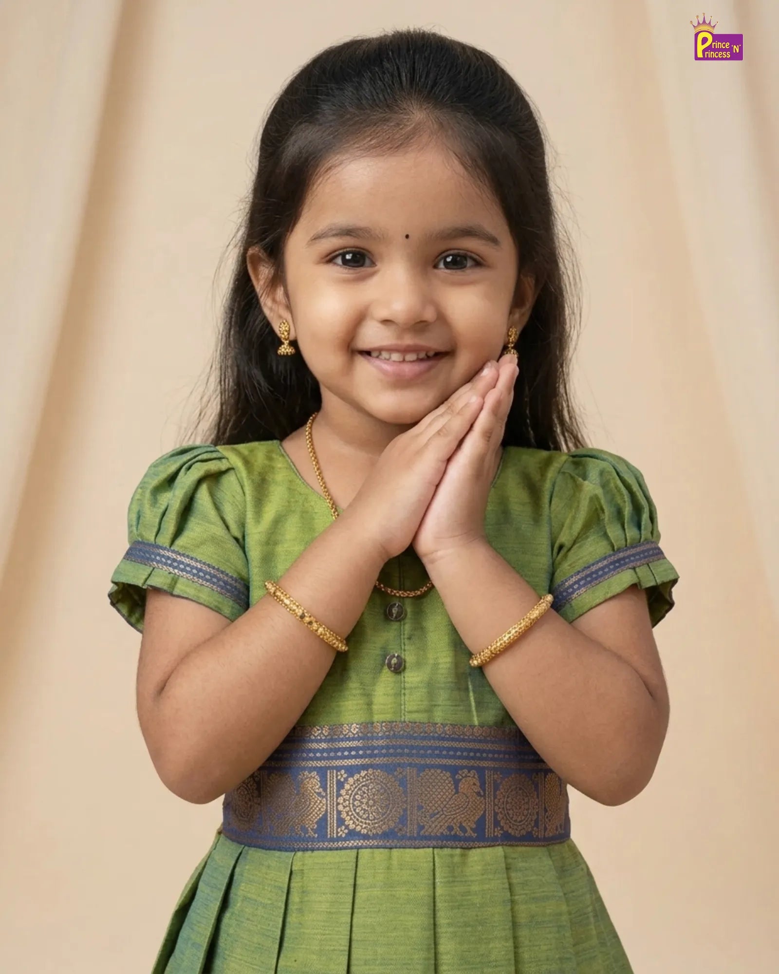 A young girl with dark hair wearing a green traditional Indian dress with gold patterns and blue trim, posing with her hands together in a 'namaste' gesture against a beige background.