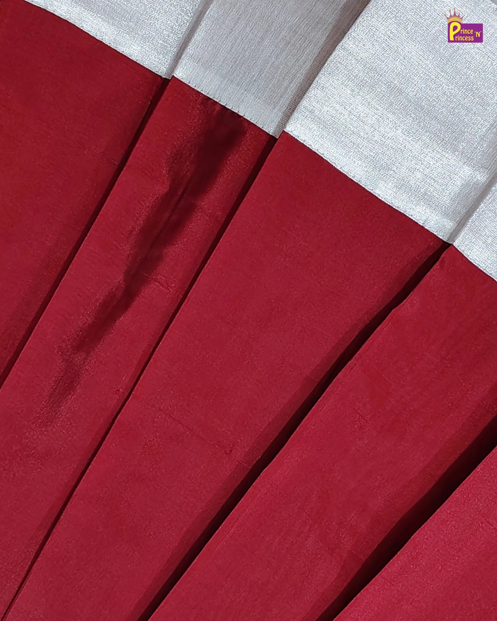 Close-up of a pleated red fabric garment, likely a skirt or drape, with a contrasting white border at the top. The fabric has a subtle sheen and is arranged in diagonal pleats.