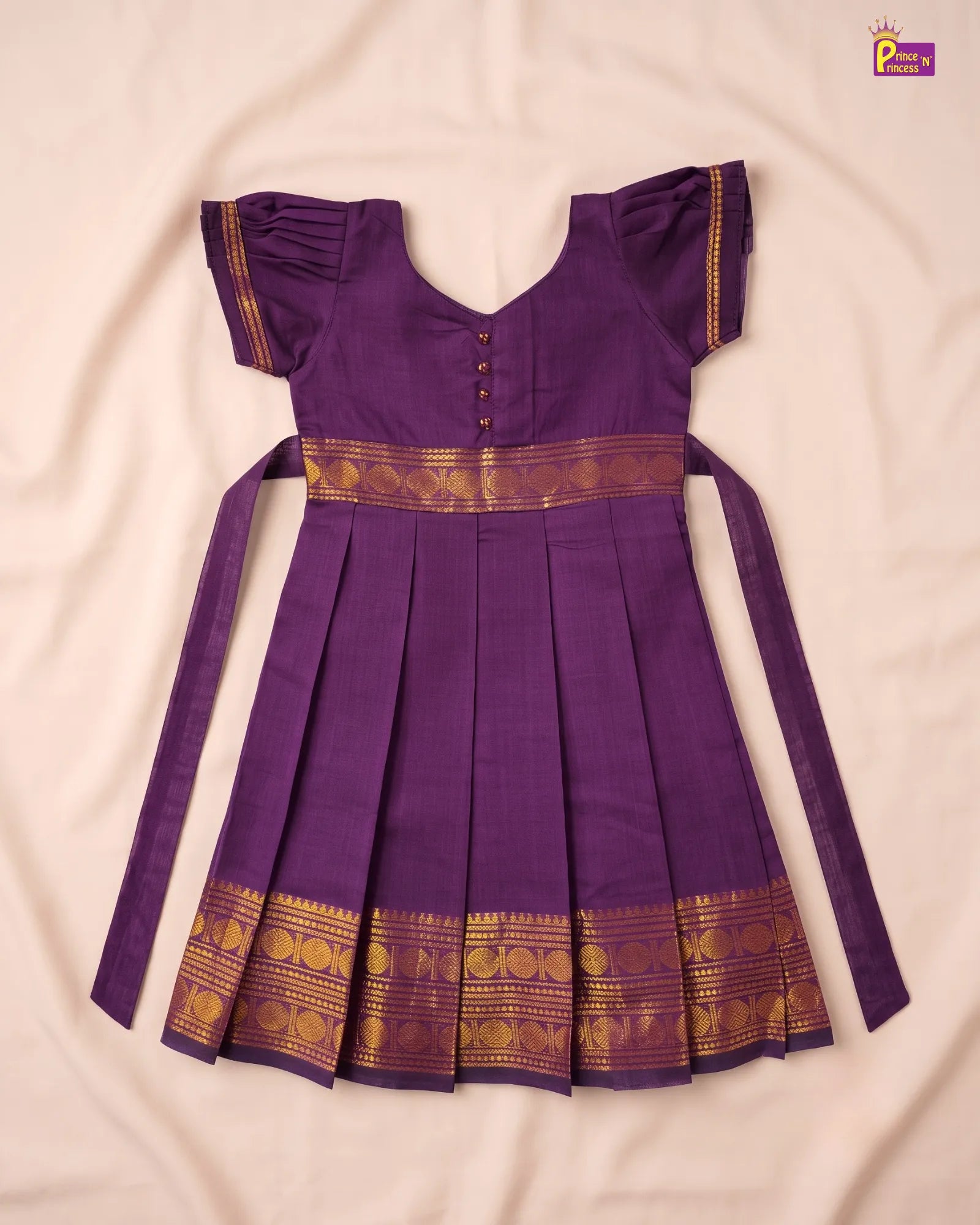 A purple traditional Indian dress with gold zari borders, laid flat on a light beige fabric background. The dress features short flutter sleeves, a V-neckline with decorative buttons, a pleated skirt, and long side ties.
