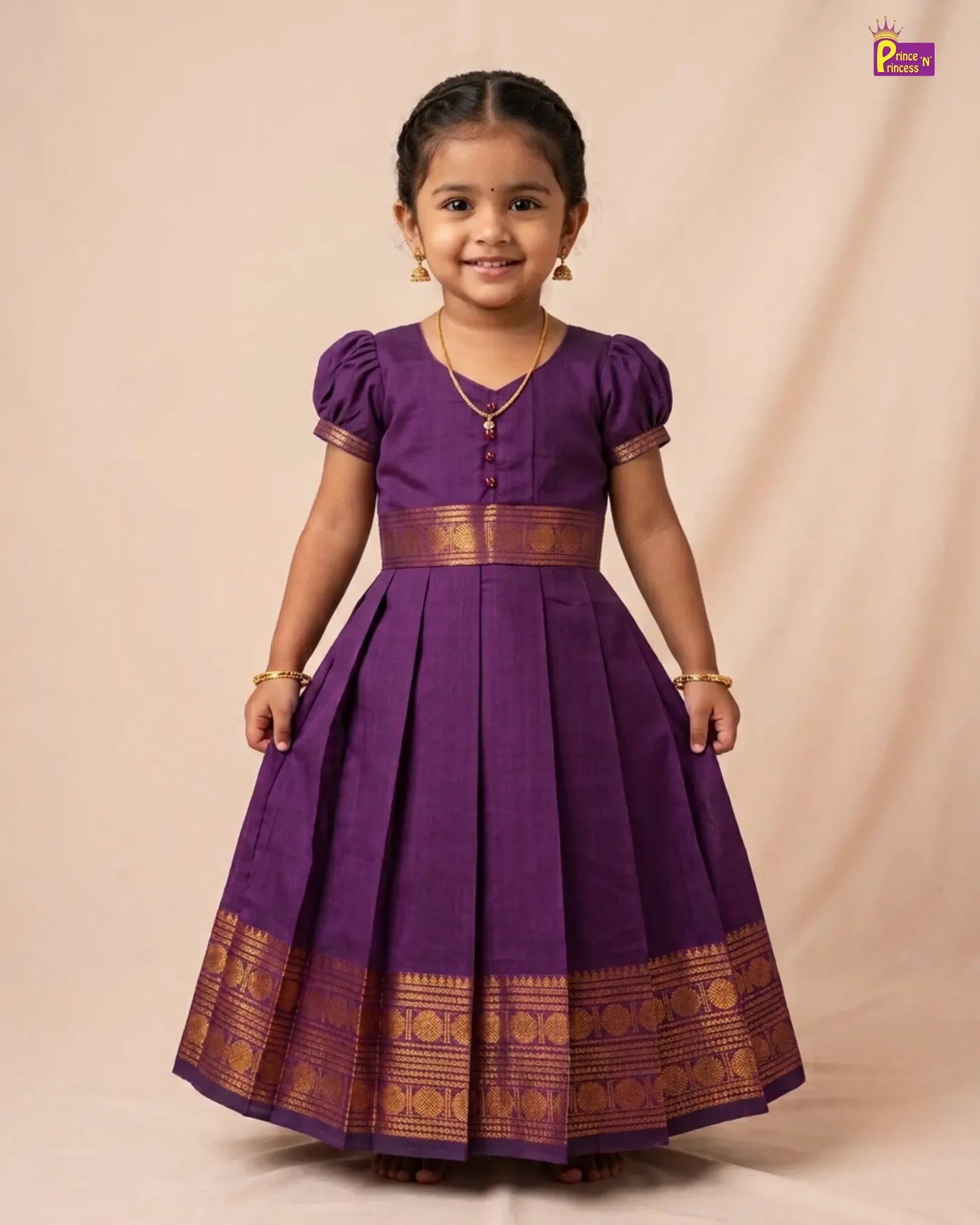 A young girl modeling a traditional purple cotton saree dress with gold zari borders and button details, standing against a beige studio backdrop.
